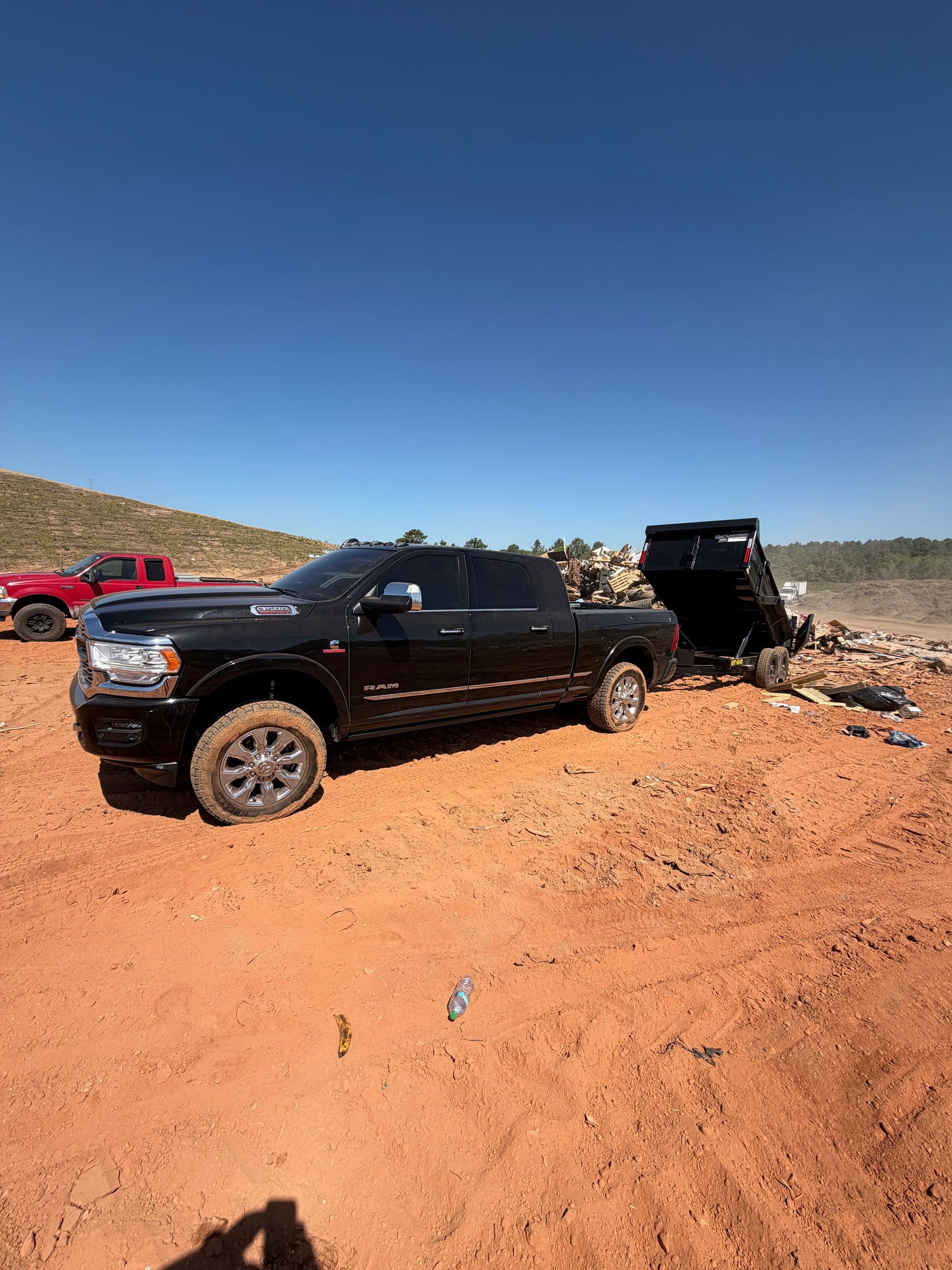 Black pickup truck towing a dump trailer on a rocky dirt road under a clear blue sky.