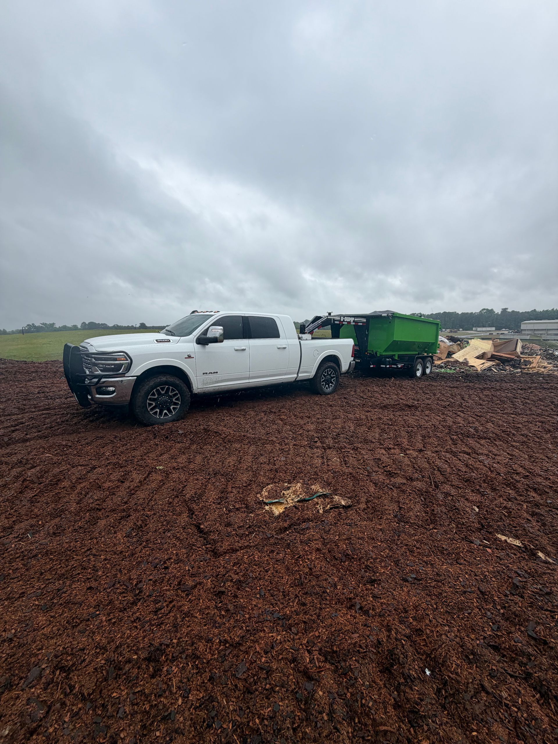 White pickup truck towing a green trailer on a muddy gravel site under cloudy skies