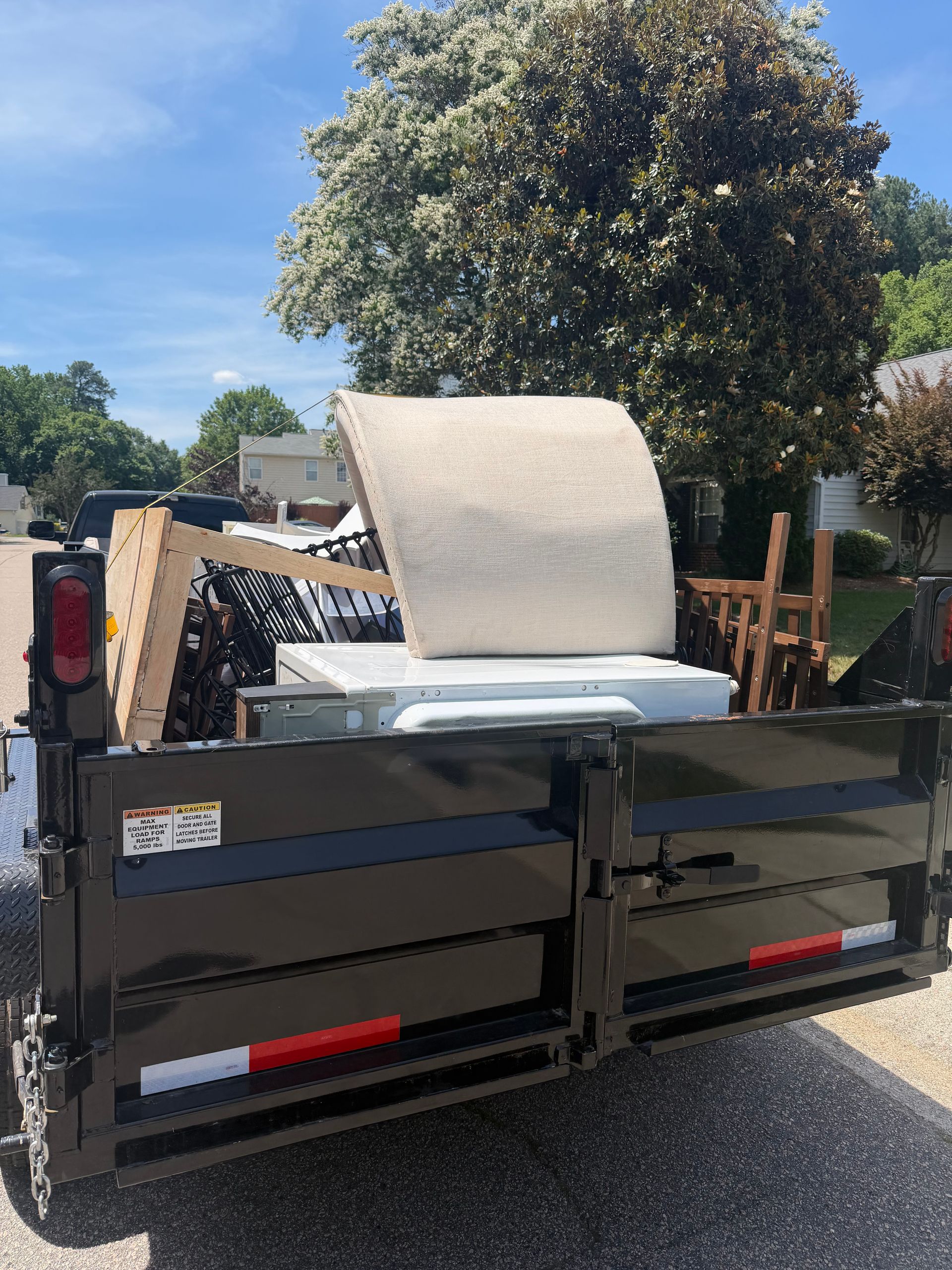 Black dumpster trailer loaded with a mattress and furniture in a sunny outdoor lot