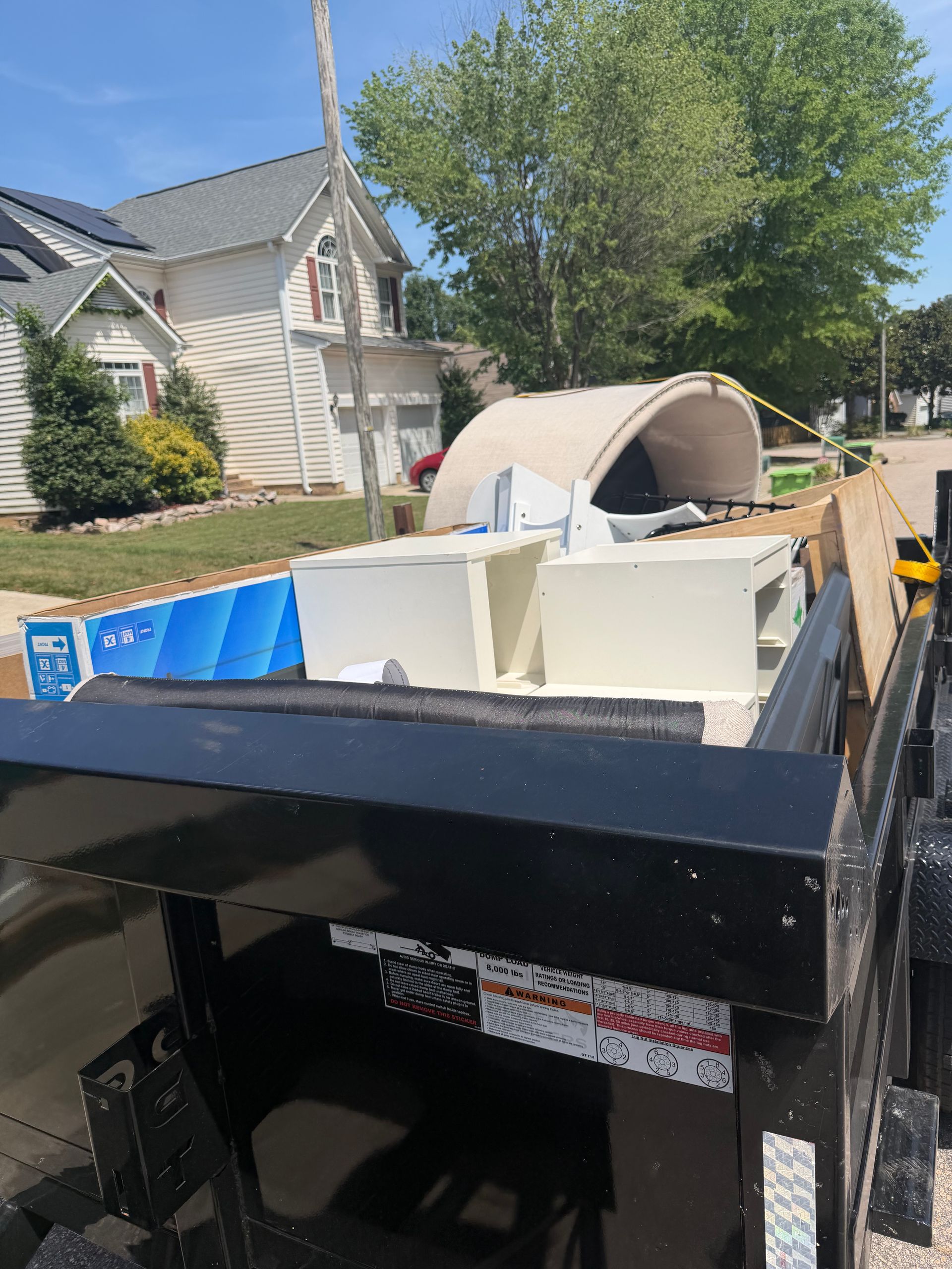 Black dumpster filled with demolition debris and furniture in a suburban driveway.