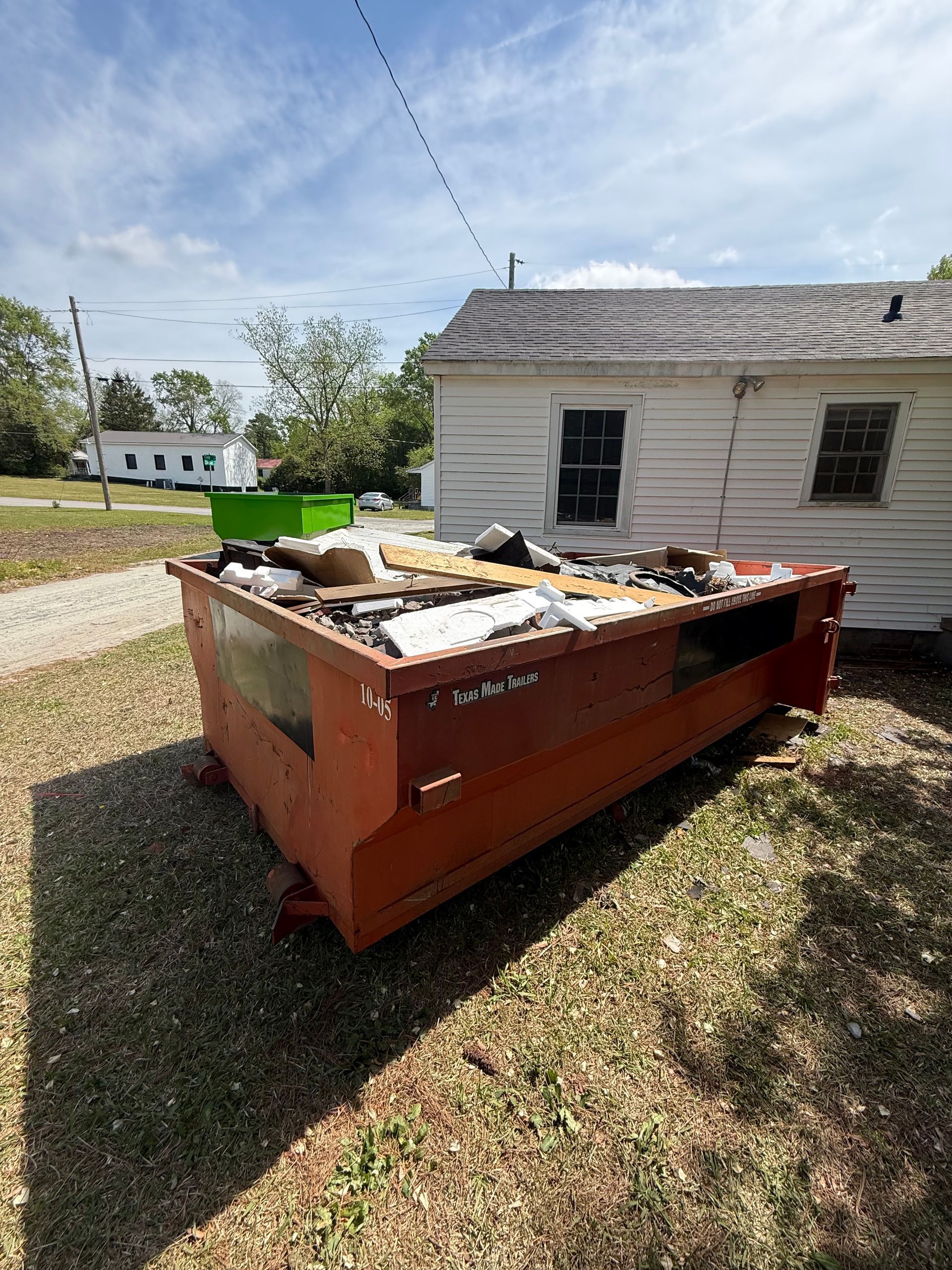 Orange dumpster filled with debris beside a small white house on a grassy lot