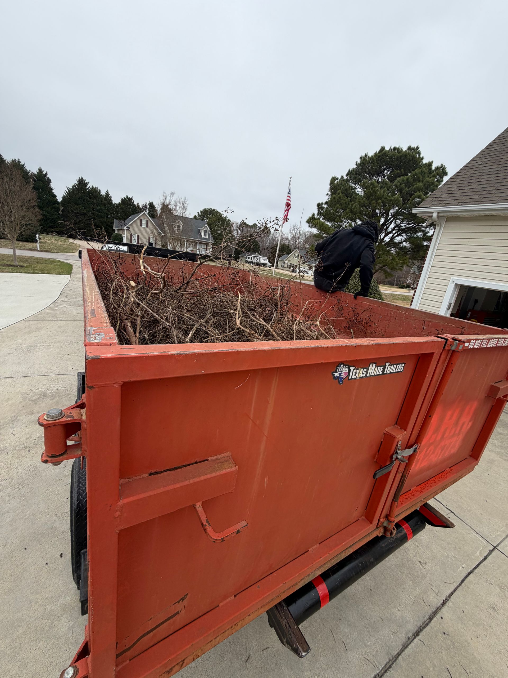 Orange yard-waste dumpster with leaf debris on a driveway; a person is loading it outdoors.