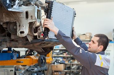 Mechanic in blue jumpsuit examining car engine, holding clipboard in a garage.