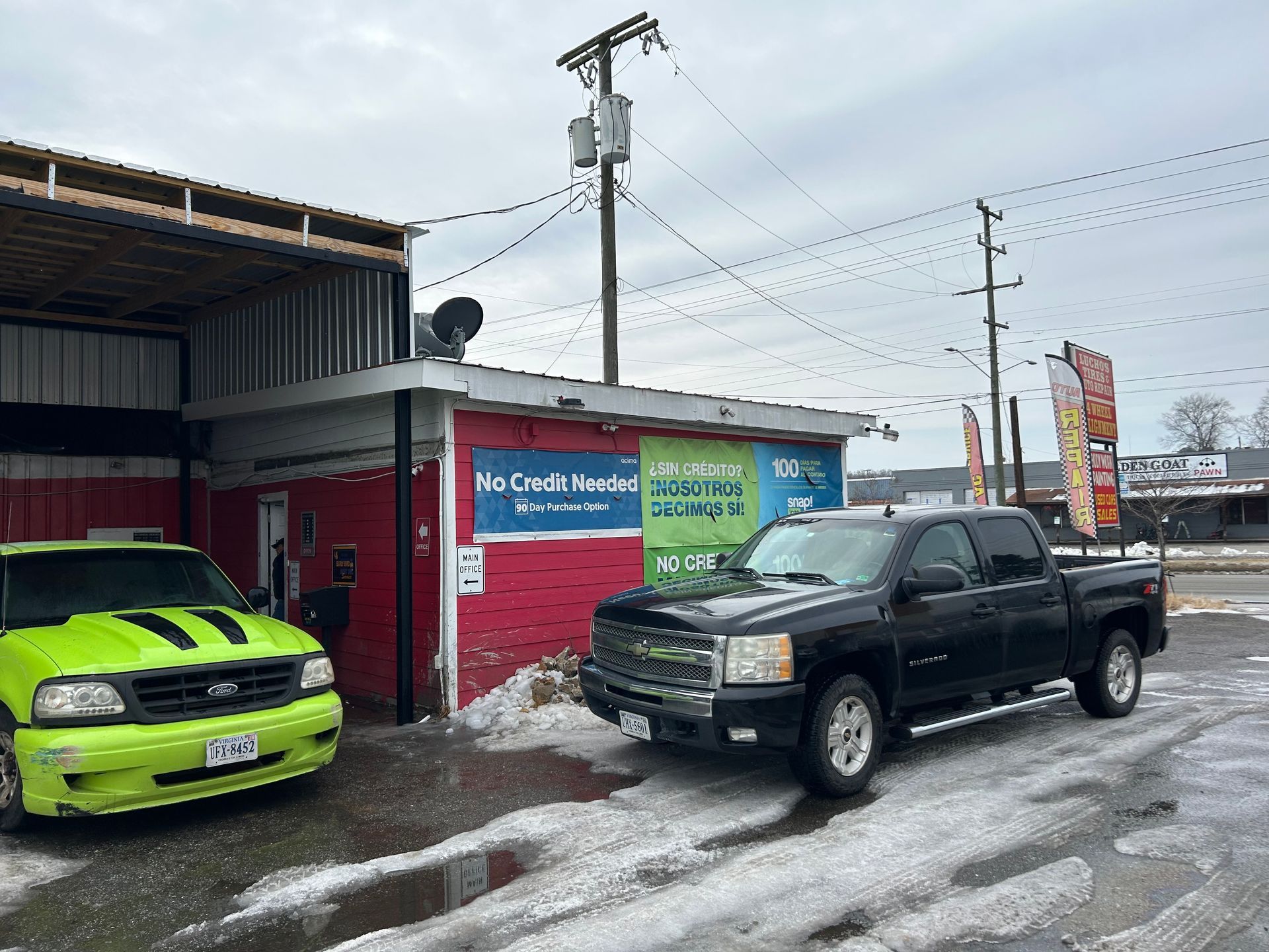 A green car and black truck parked outside a red and white auto repair shop on a cloudy day.