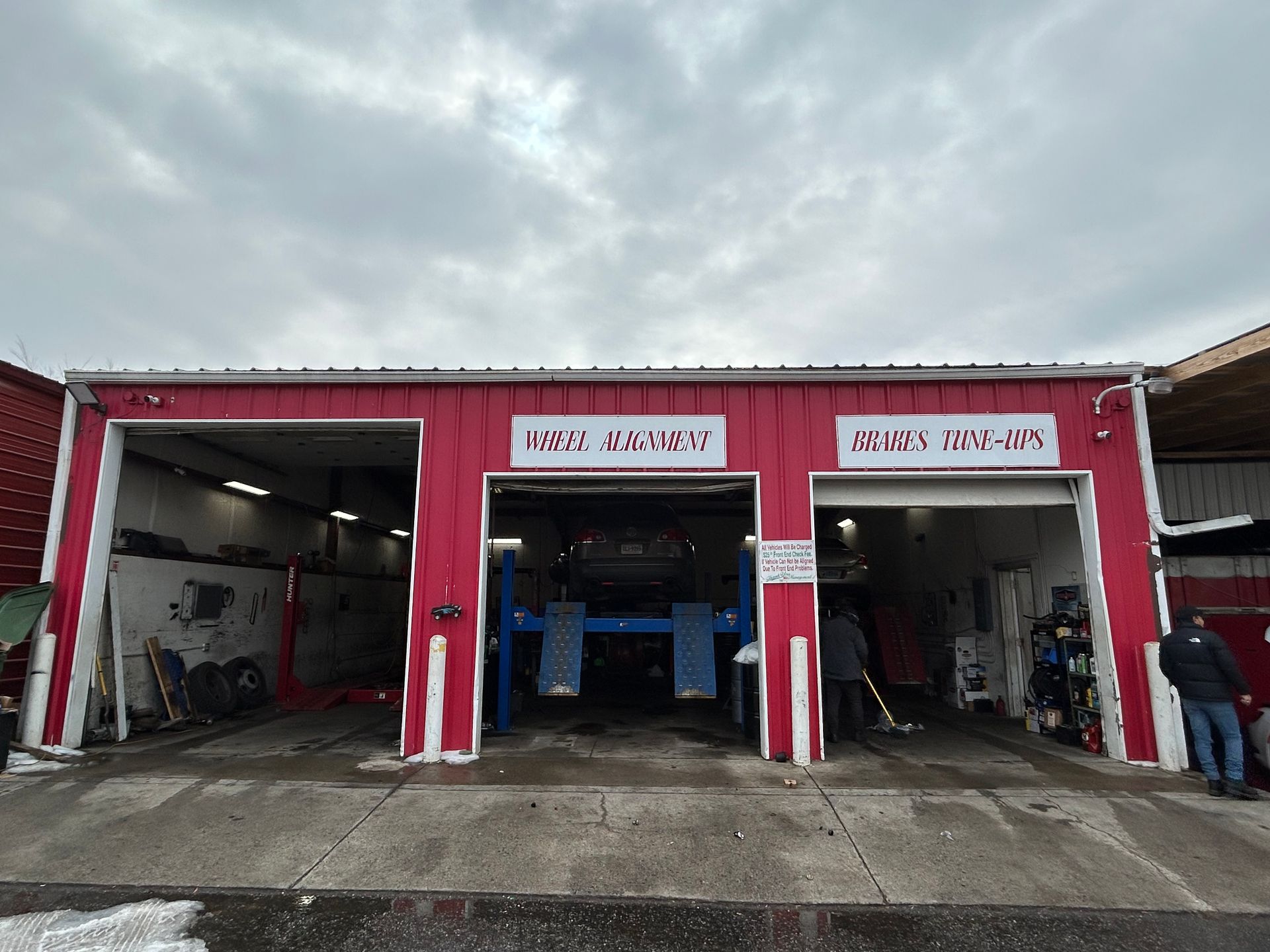 Red auto repair shop with open bays; a car is on a lift. People are working inside. Cloudy sky.