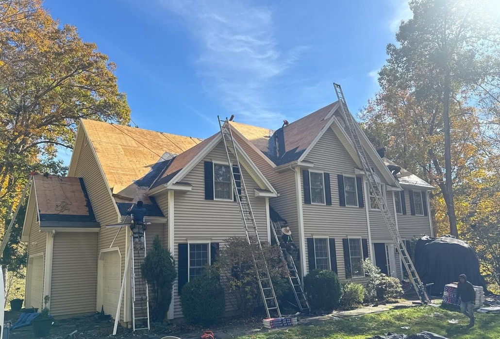 Large beige house with dark shutters, steep gables, and ladders under a sunny blue sky