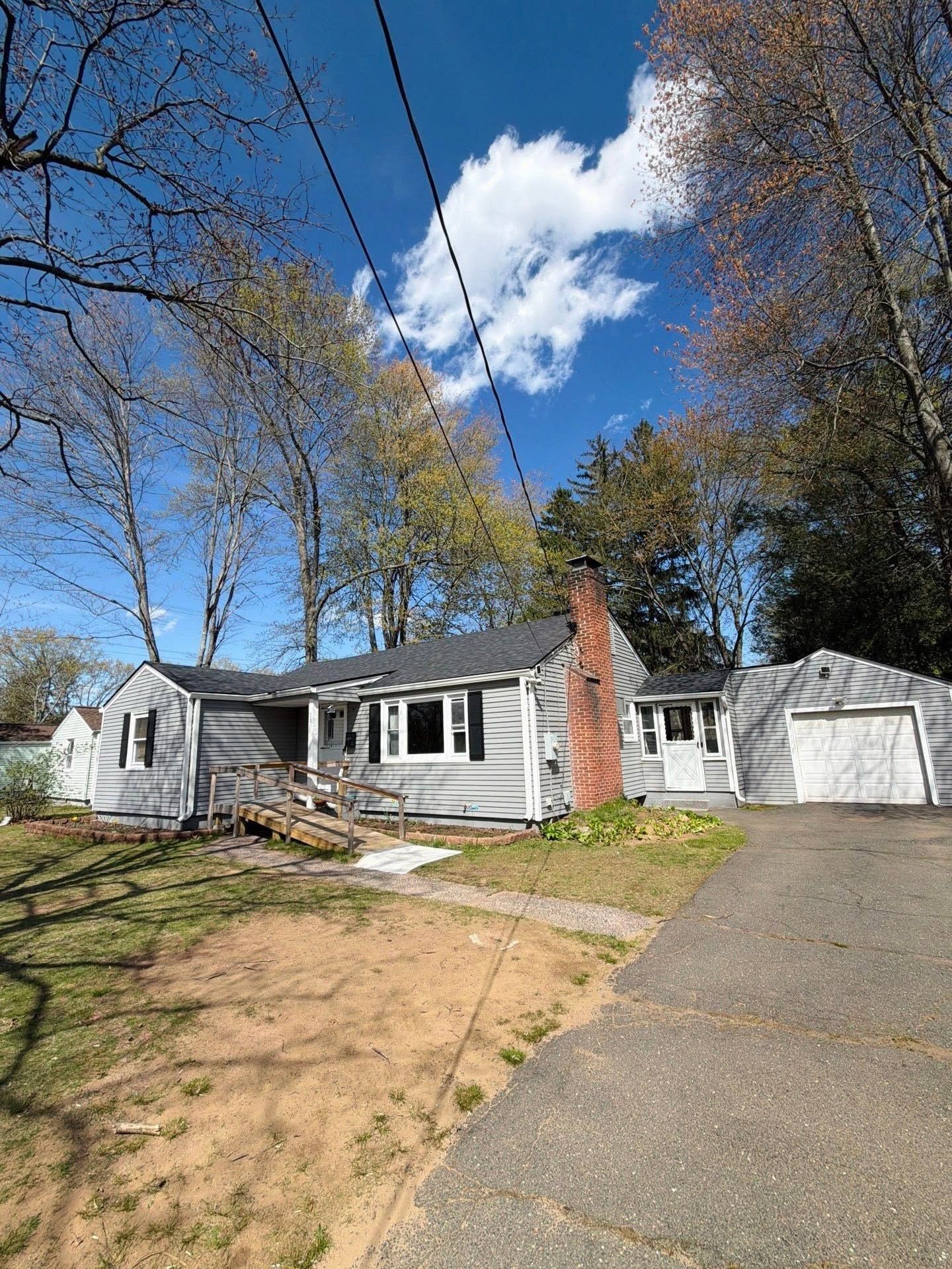Small white house with brick chimney and detached garage on a sunny suburban lot.