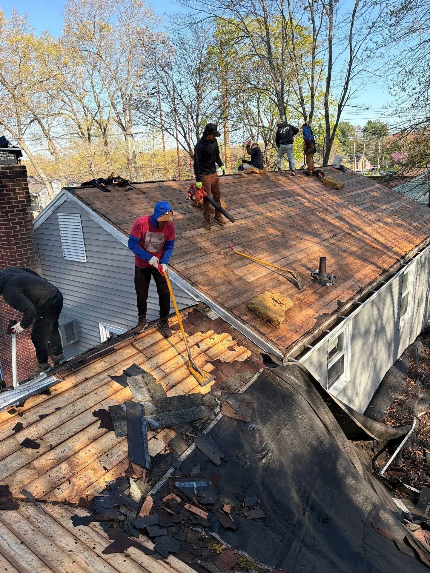 Workers replacing shingles on a steep roof beside trees and a brick chimney.