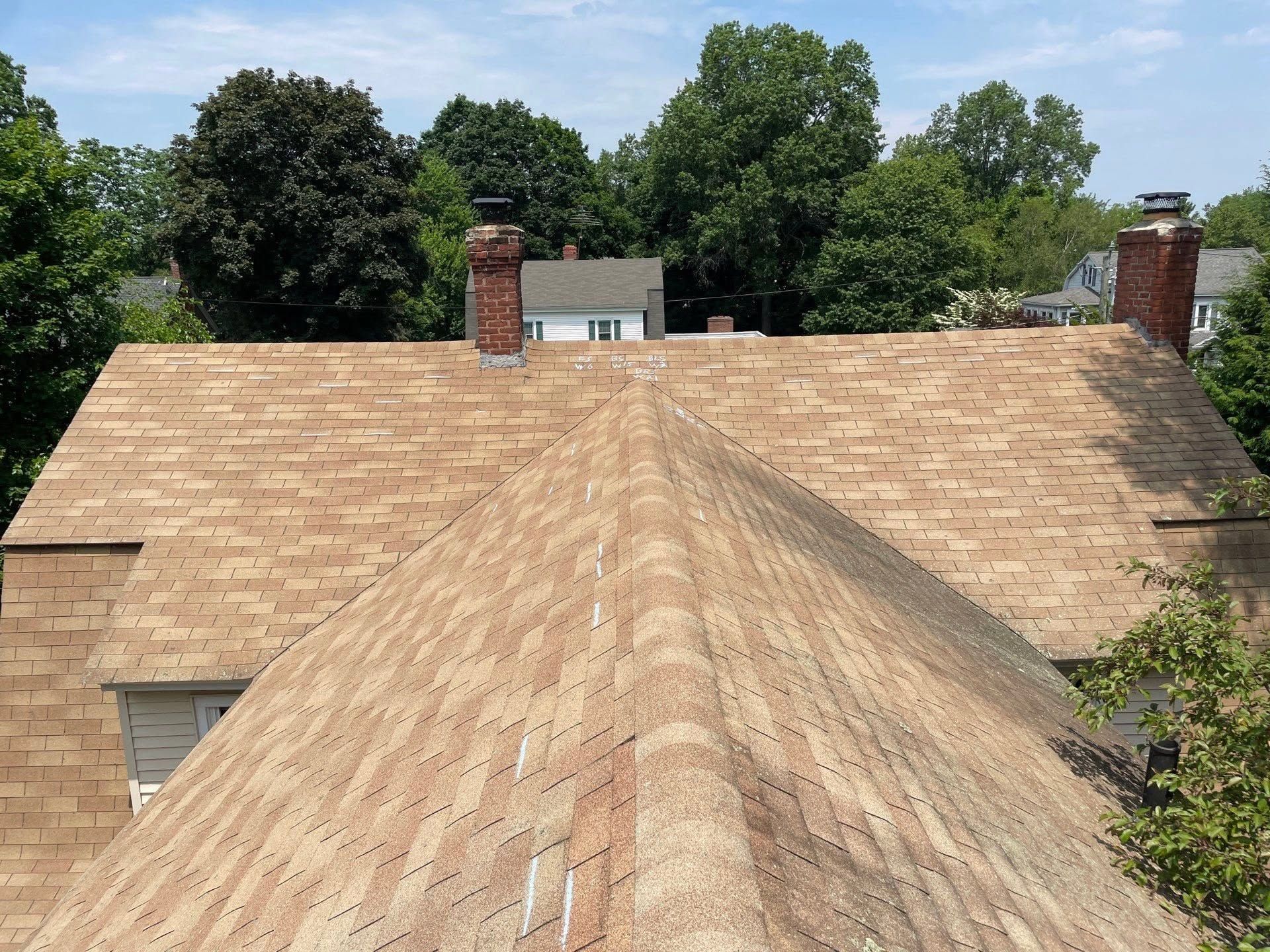 Tan shingled house roof with two brick chimneys, viewed from above with trees in the background