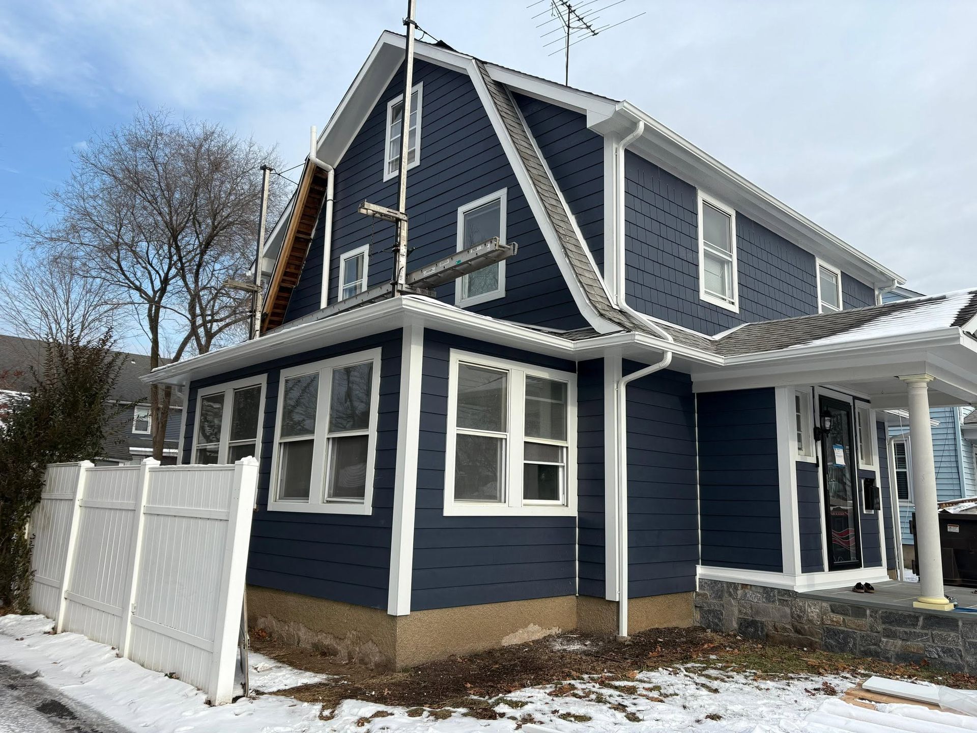 Blue two-story house with white trim, porch, and fence on a snowy winter day