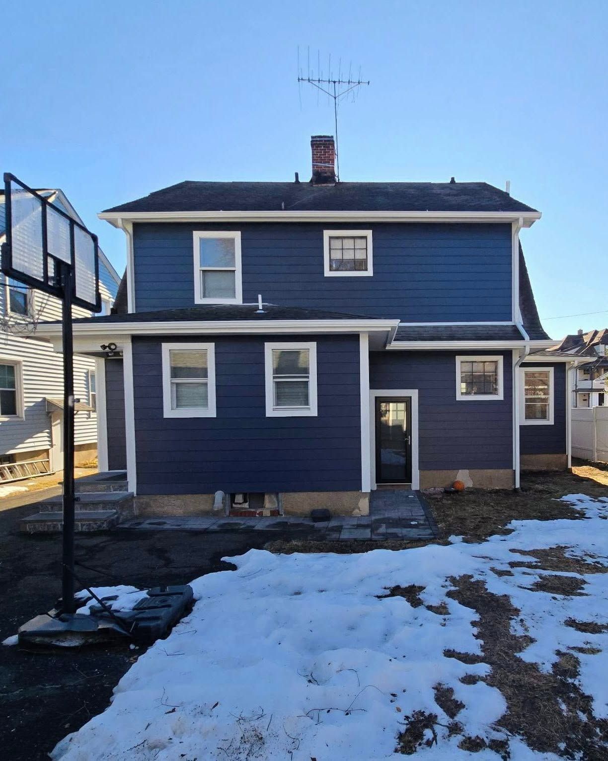 Blue two-story house with white trim and a patchy snow-covered yard on a sunny day