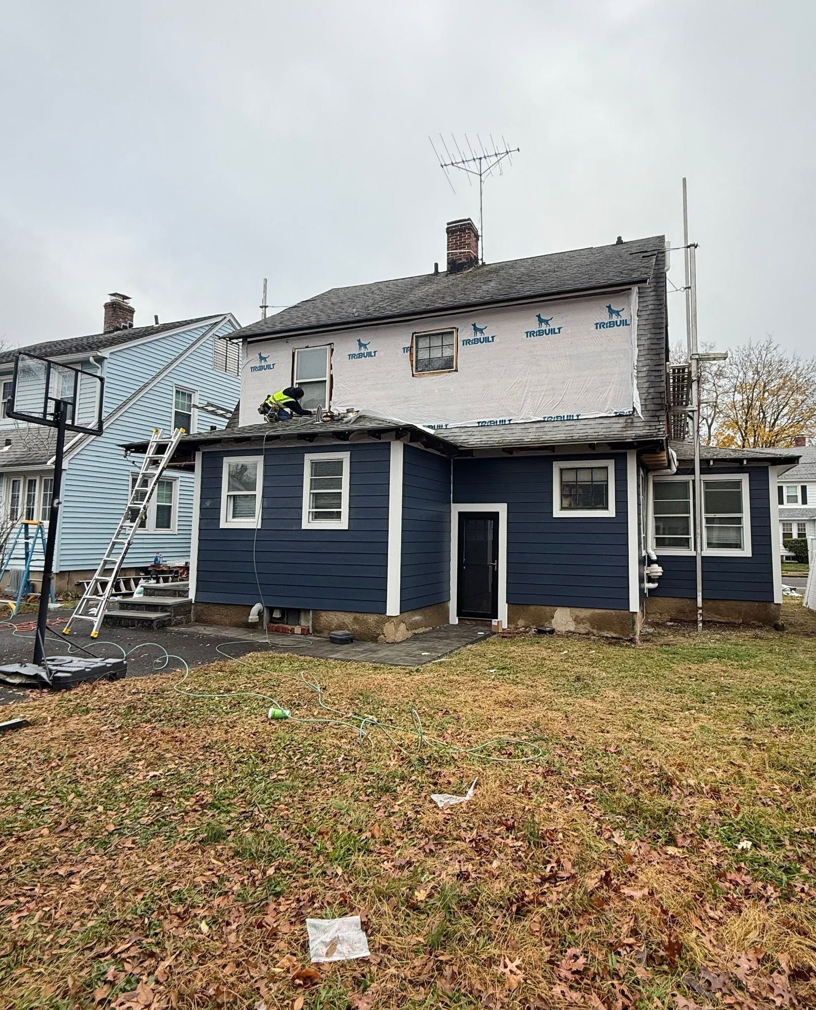 Damaged two-story house with blue siding, exposed roof, and fallen debris in a wet yard.
