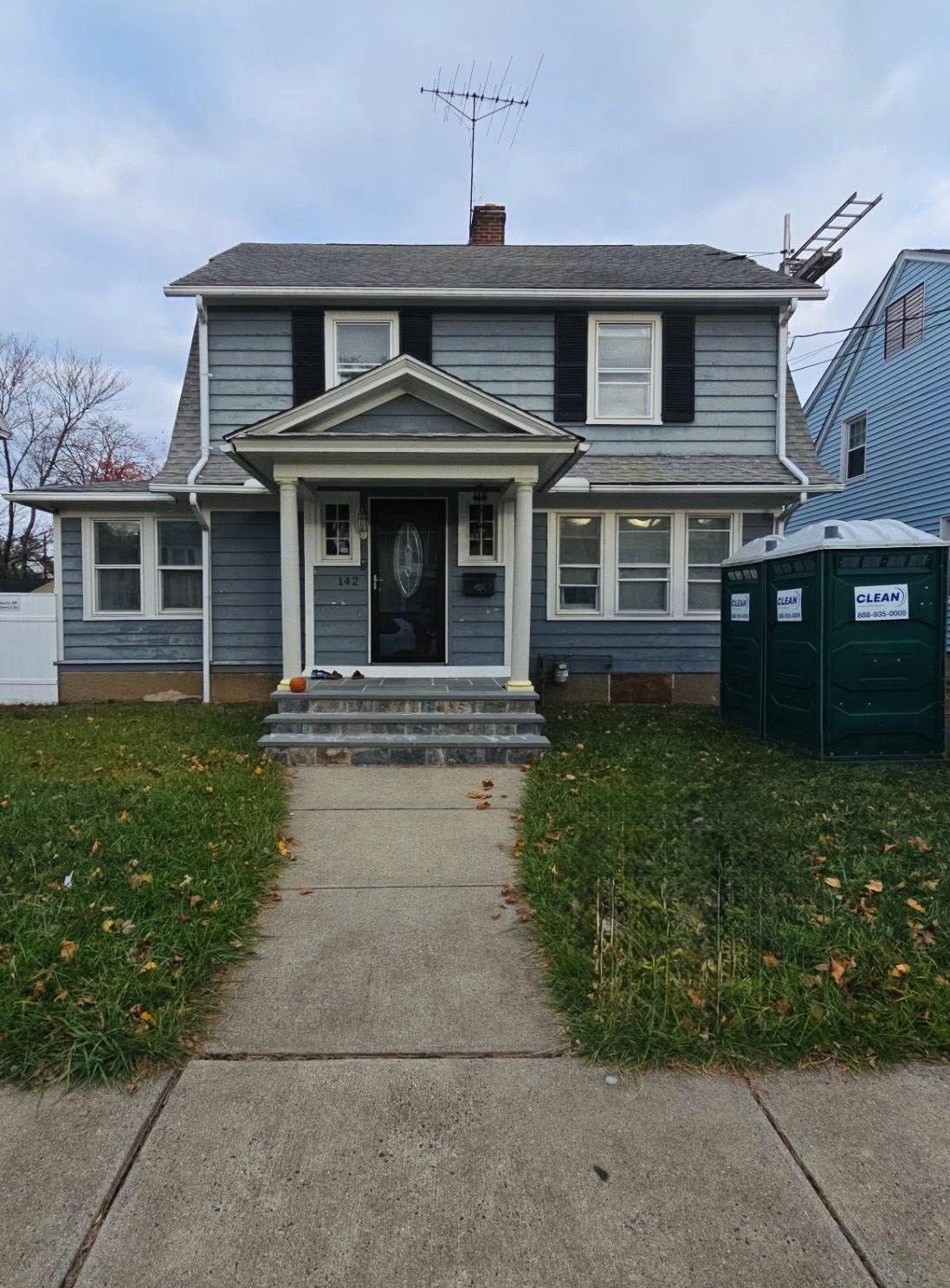 Two-story gray house with a front porch, steps, lawn, and sidewalk, with a green portable toilet on the right.