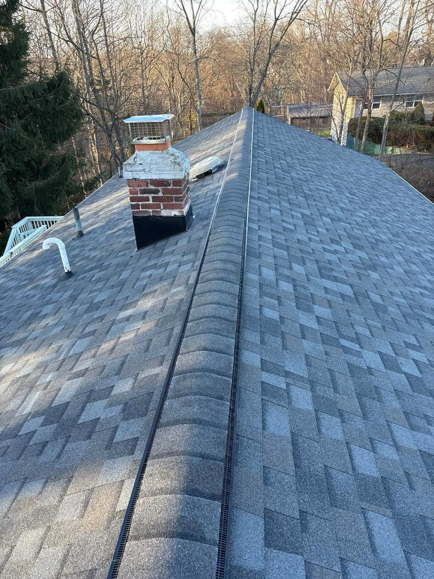 Steep gray shingle roof with brick chimney and white vent pipes, trees and neighboring houses in background