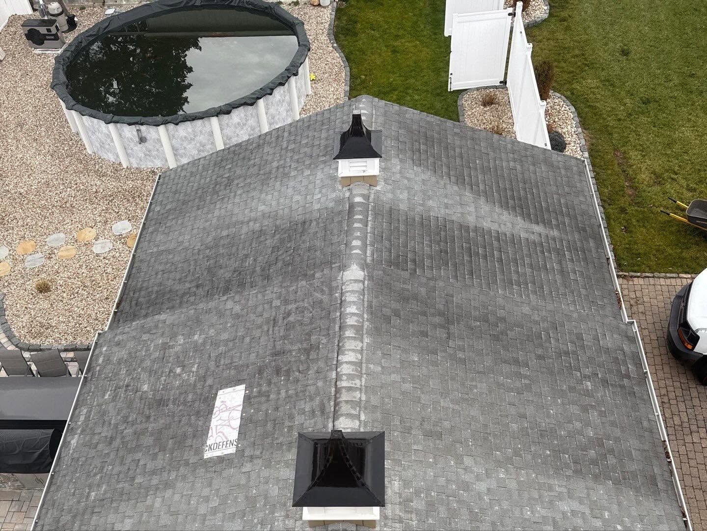 Aerial view of a gray roof with two vents beside a circular pool and yard.