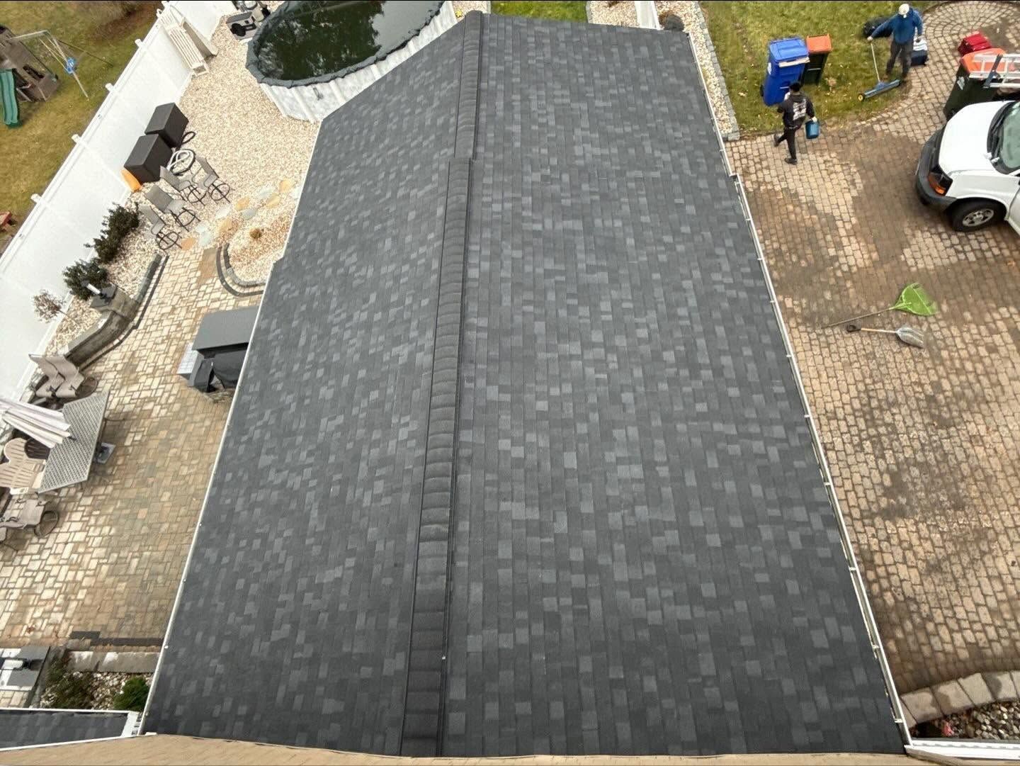 Aerial view of a dark shingled roof with workers and a white truck in a driveway below