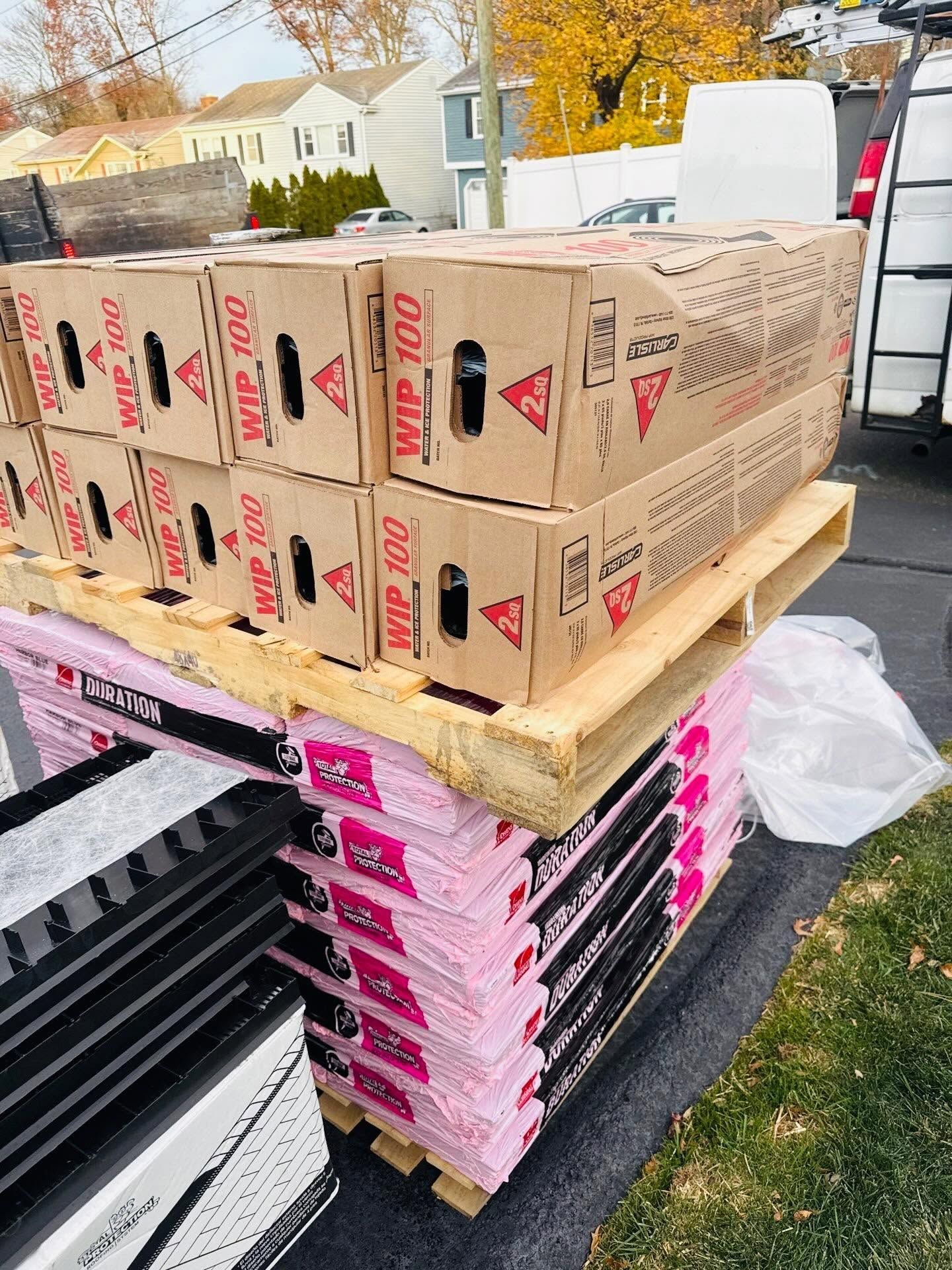 Stacked construction material boxes on wooden pallets outdoors beside a trailer and grassy yard.