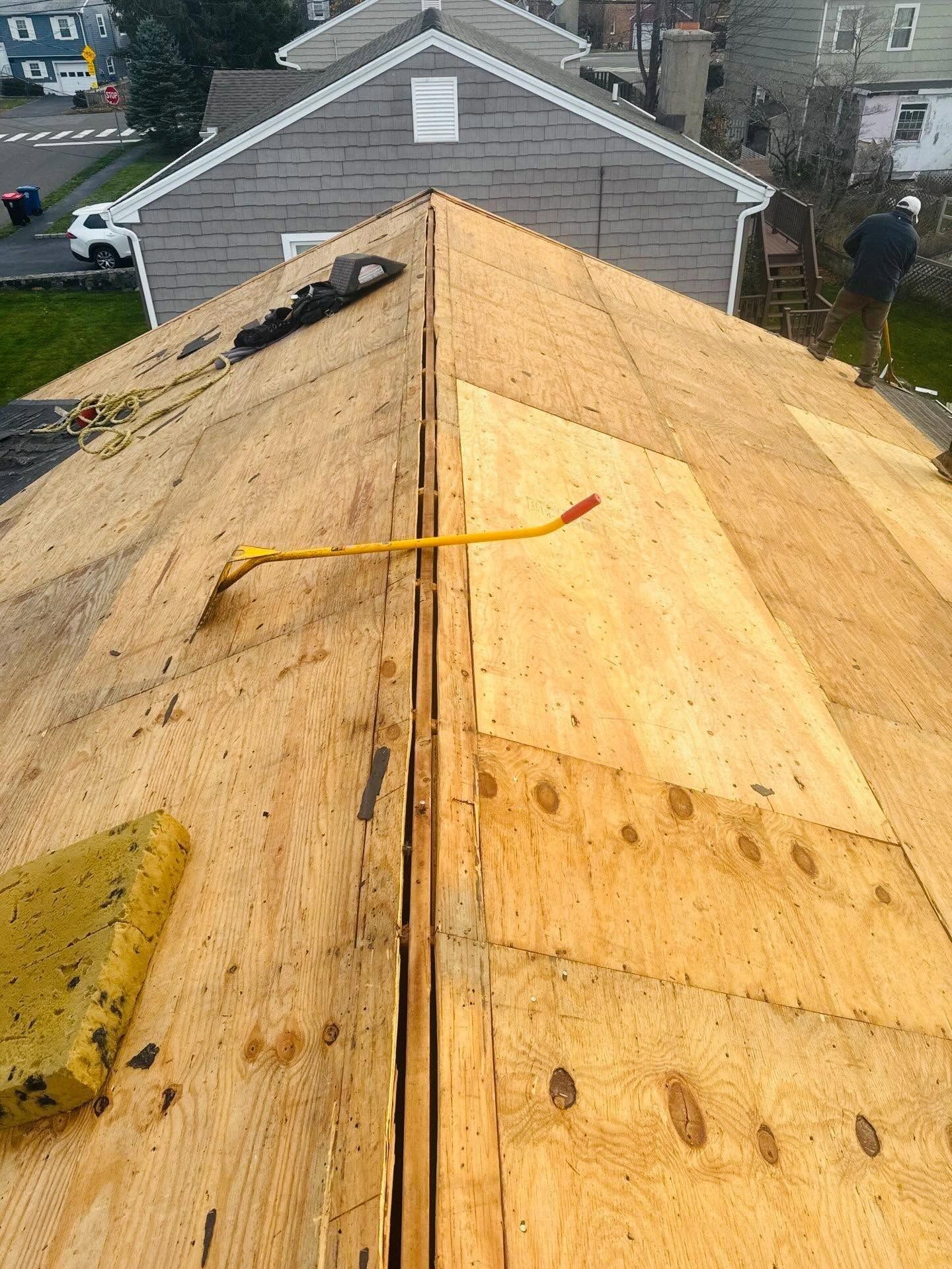 Roof under repair with plywood sheathing, yellow strap, and a neighboring house in the background.