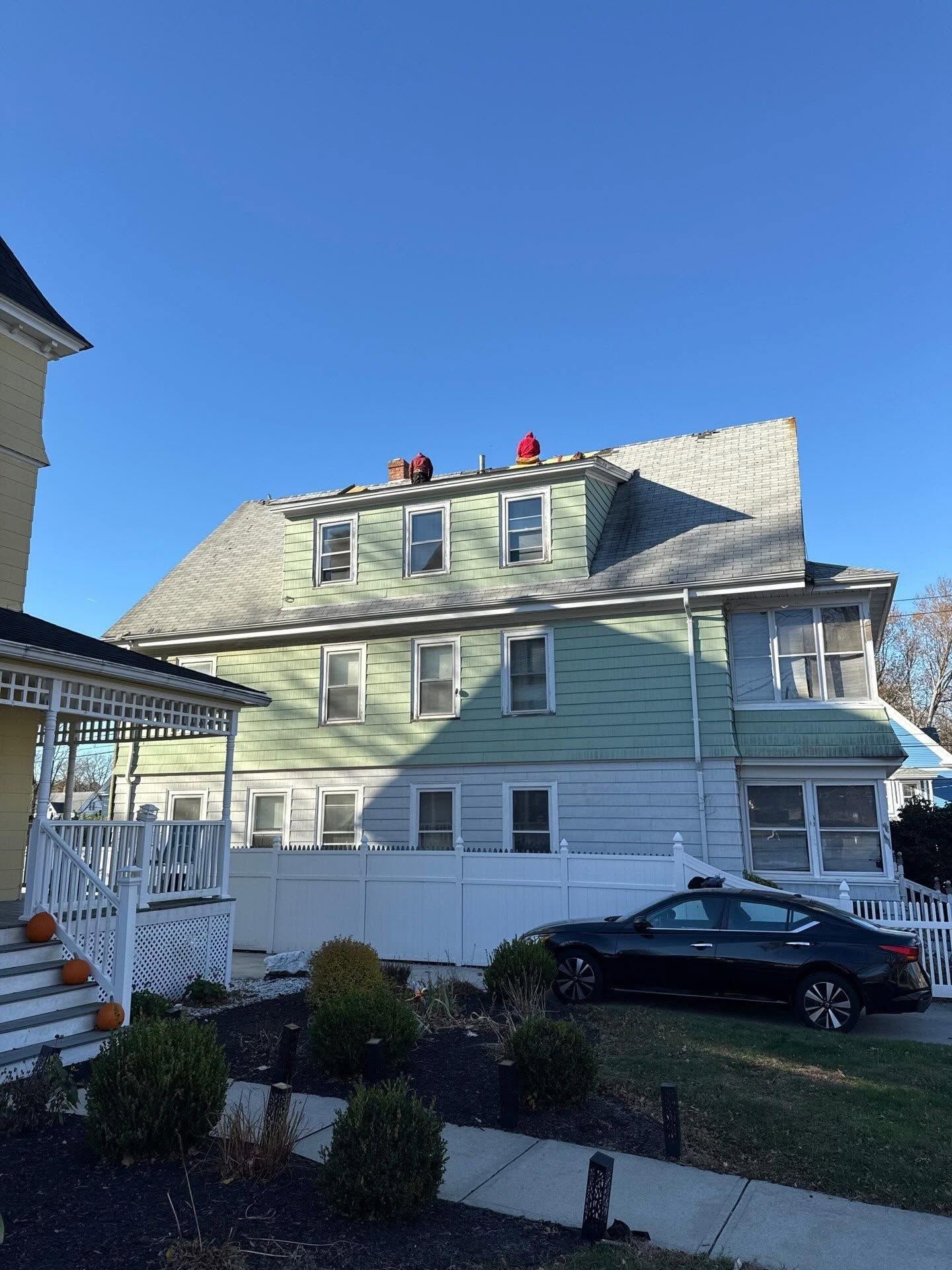 Light green house with white trim, dormer windows, white fence, and a black car parked in front