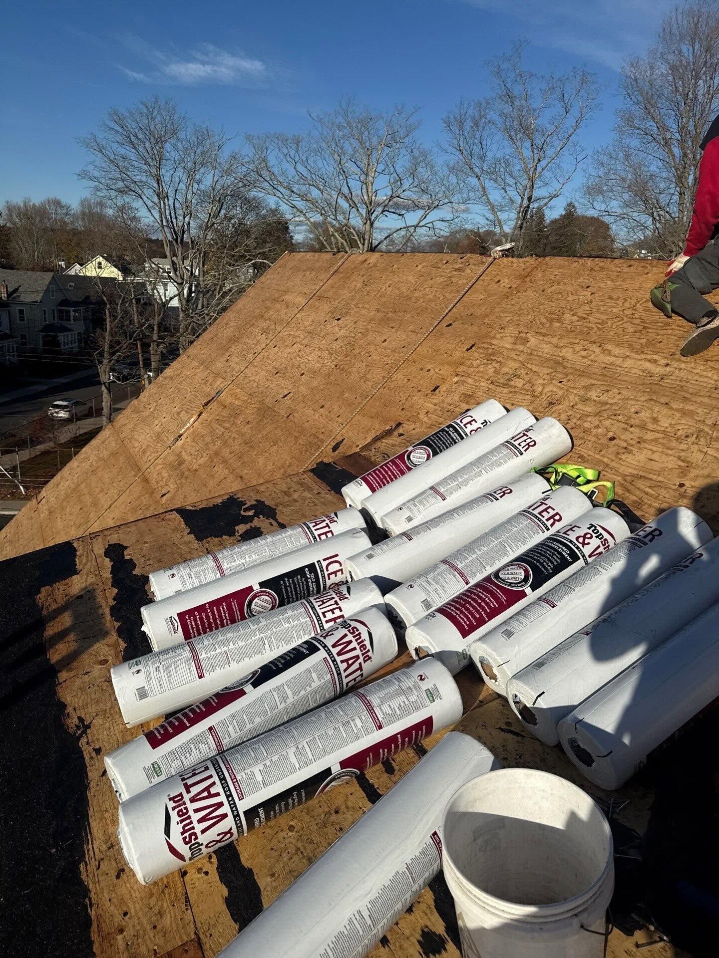 Roof under construction with rolled roofing materials and a bucket on plywood decking.