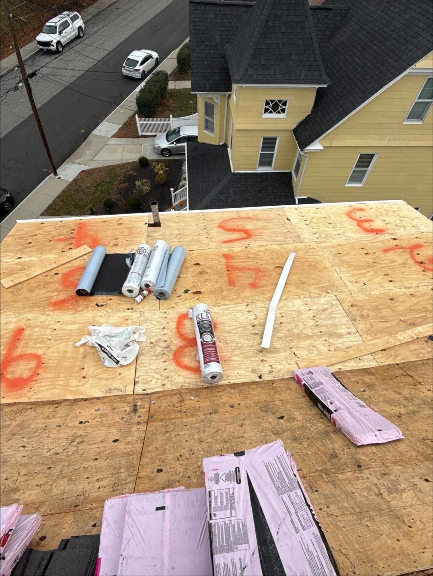 Roof under construction with rolls of roofing material and tools laid on plywood decking above houses.