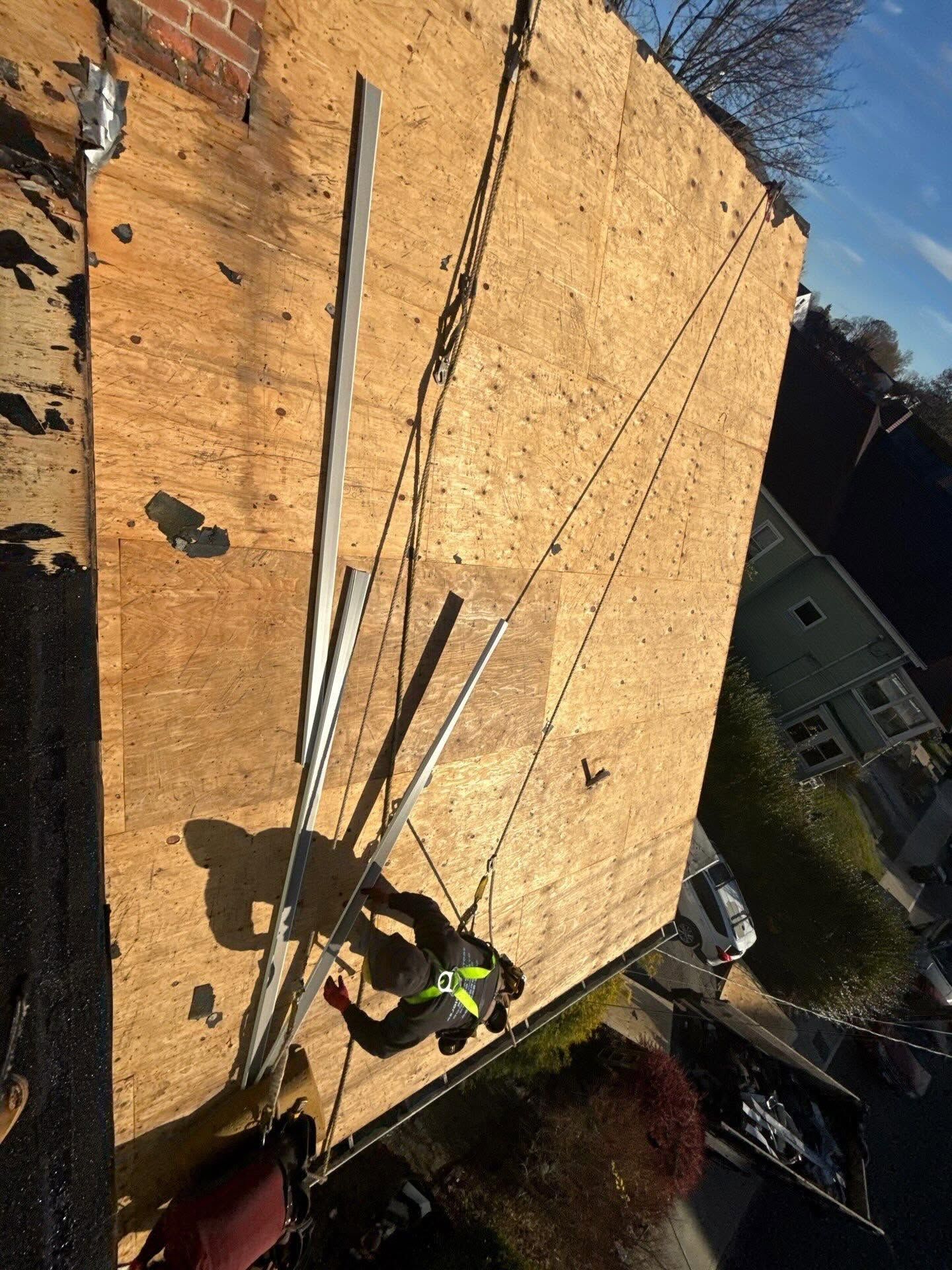 Worker on a plywood roof, securing long white beams near a house in a suburban neighborhood.