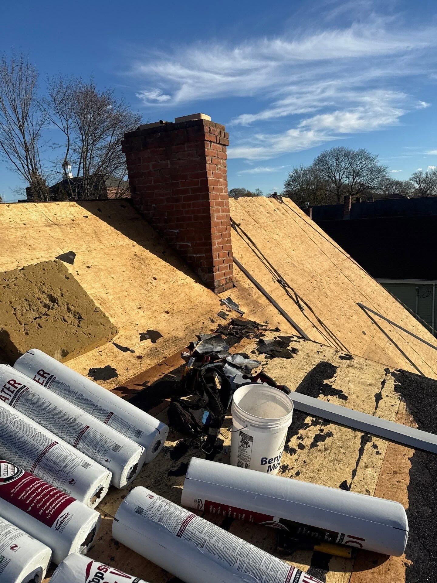 Rooftop under construction with plywood sheathing, a brick chimney, and stacks of roofing materials.