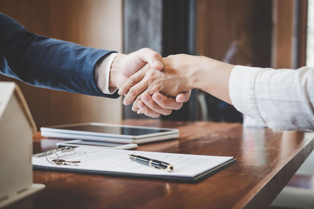 Two people shaking hands across a wooden desk with a clipboard and pen in the foreground
