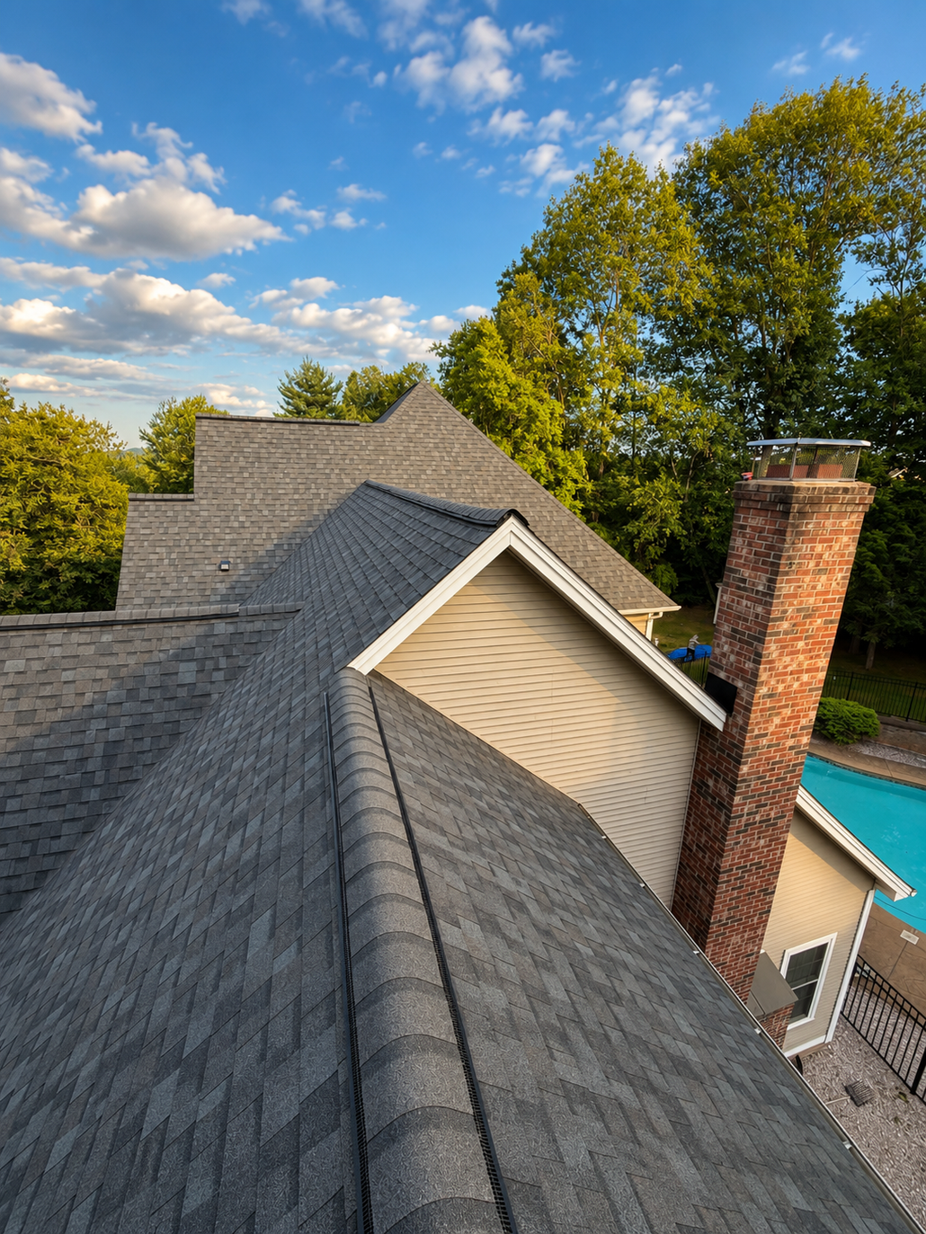 Rooftop shingles, brick chimney, and blue sky with clouds above a tree-lined suburban home