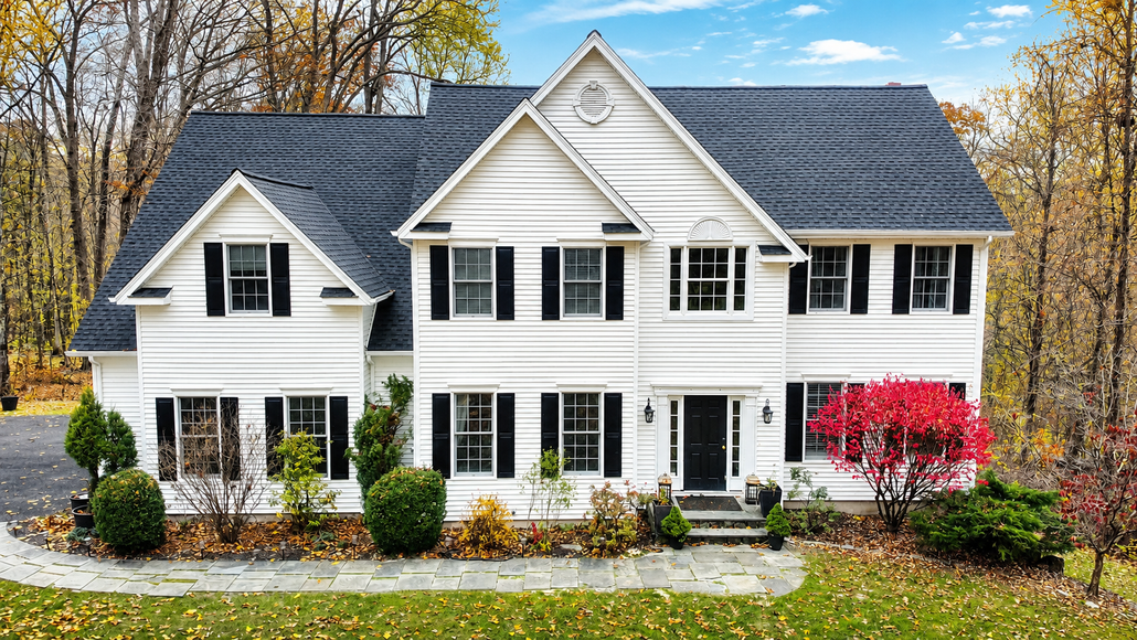 Large white two-story house with black shutters and a dark roof, surrounded by autumn trees and landscaping