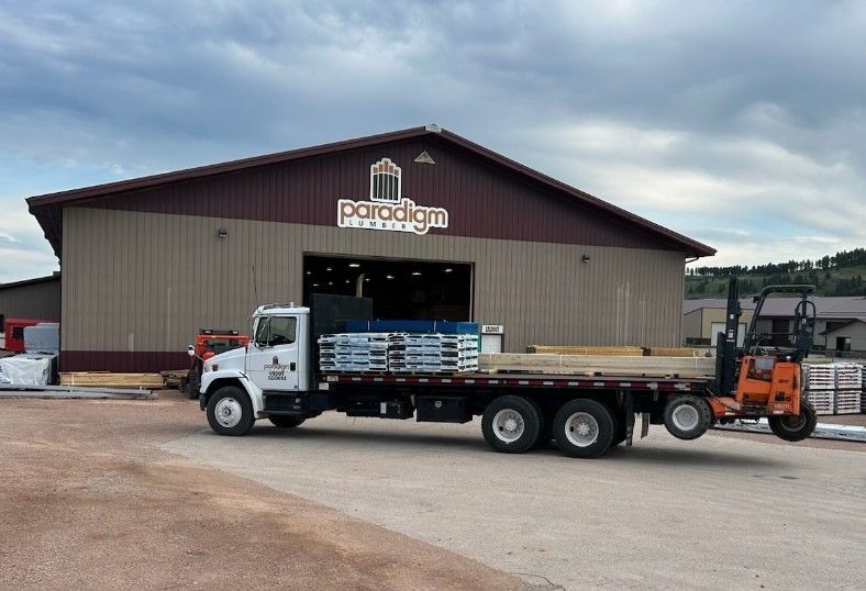 Truck loading lumber at Palladium Lumber building. Orange forklift, brown and tan building, gray truck.