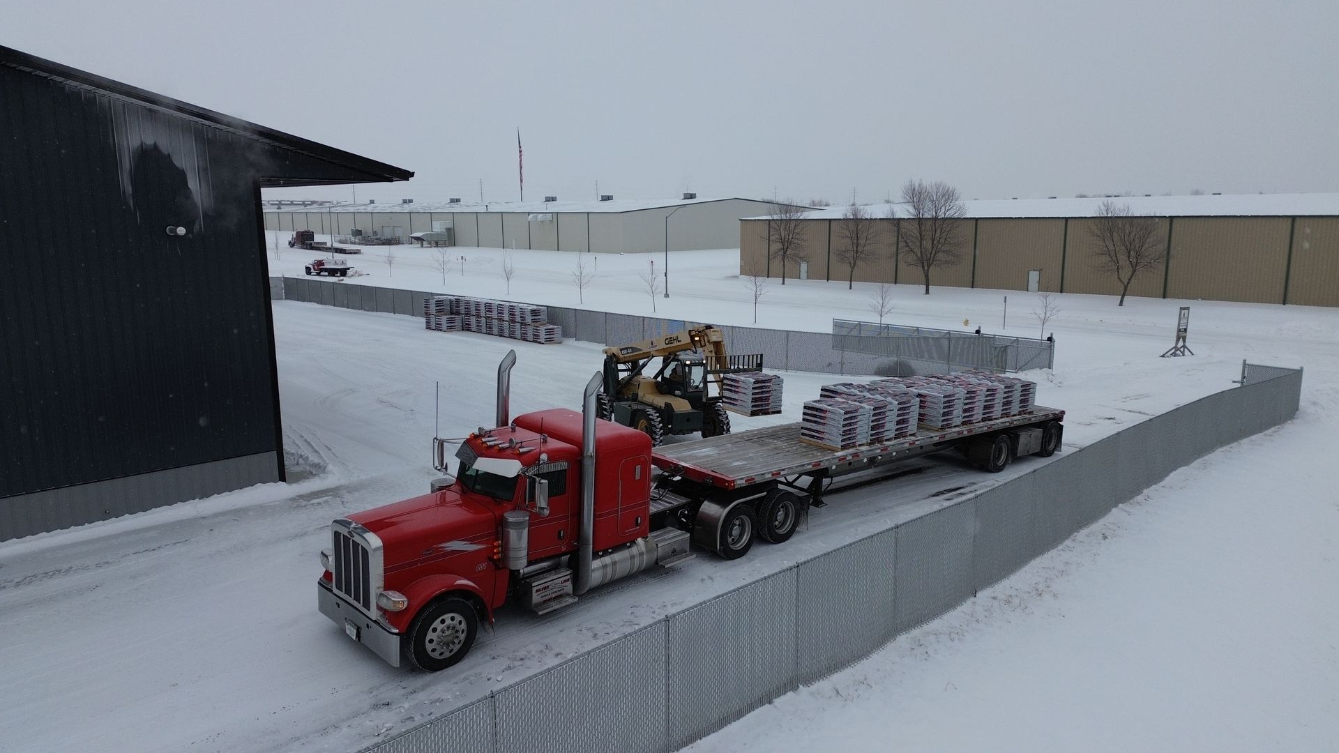 Red semi-truck loaded with materials outside a warehouse in a snowy area. A forklift loads more materials.