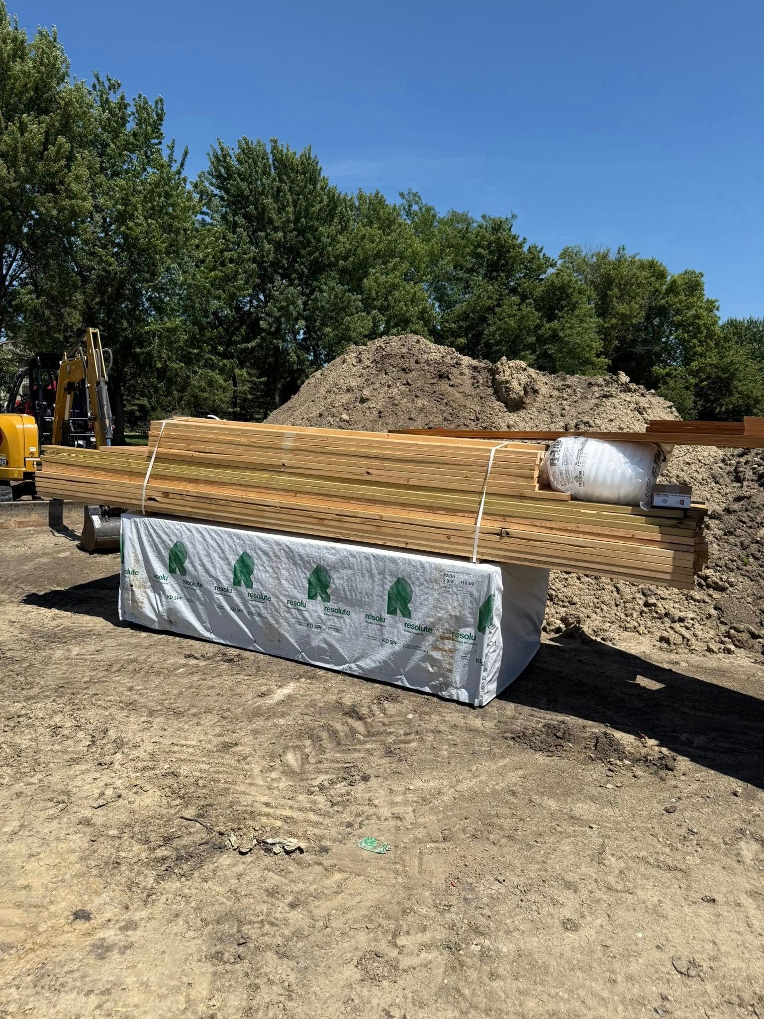 Lumber bundles wrapped, stacked outdoors on dirt, with trees and a blue sky in the background.