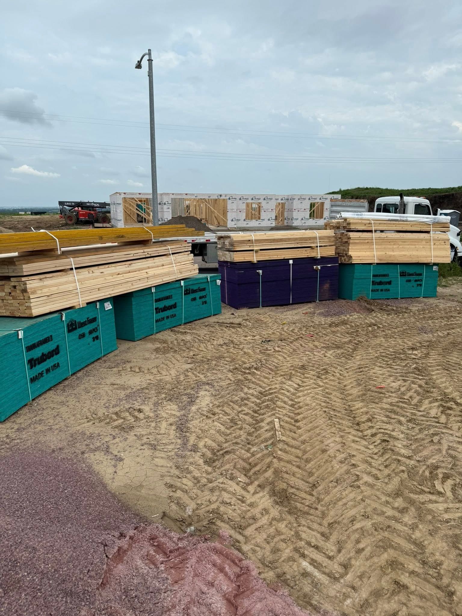 Construction site with stacks of lumber in various colors. Building frame visible in the background.