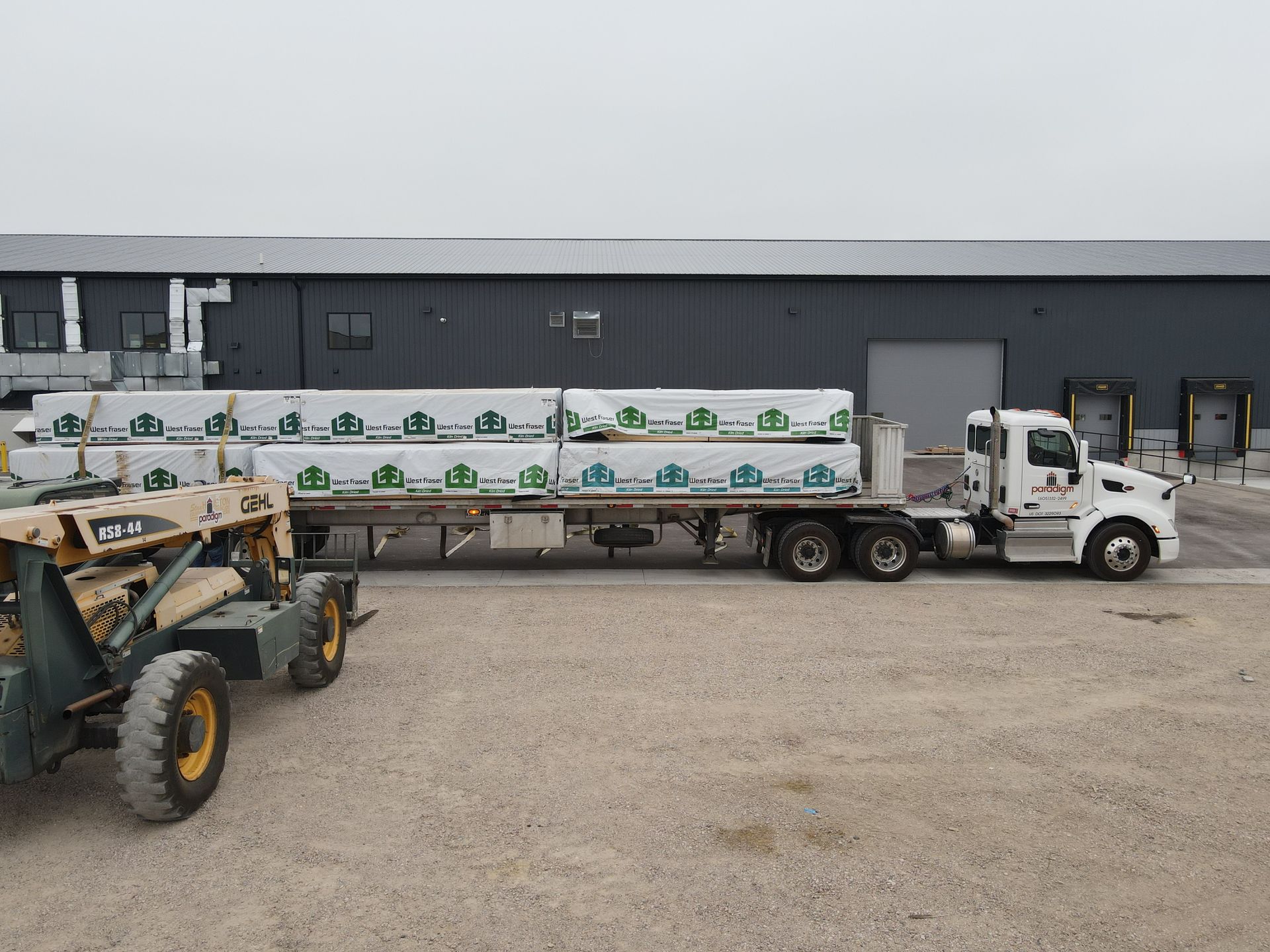 Flatbed truck carrying lumber, parked outside a warehouse. A forklift sits nearby.
