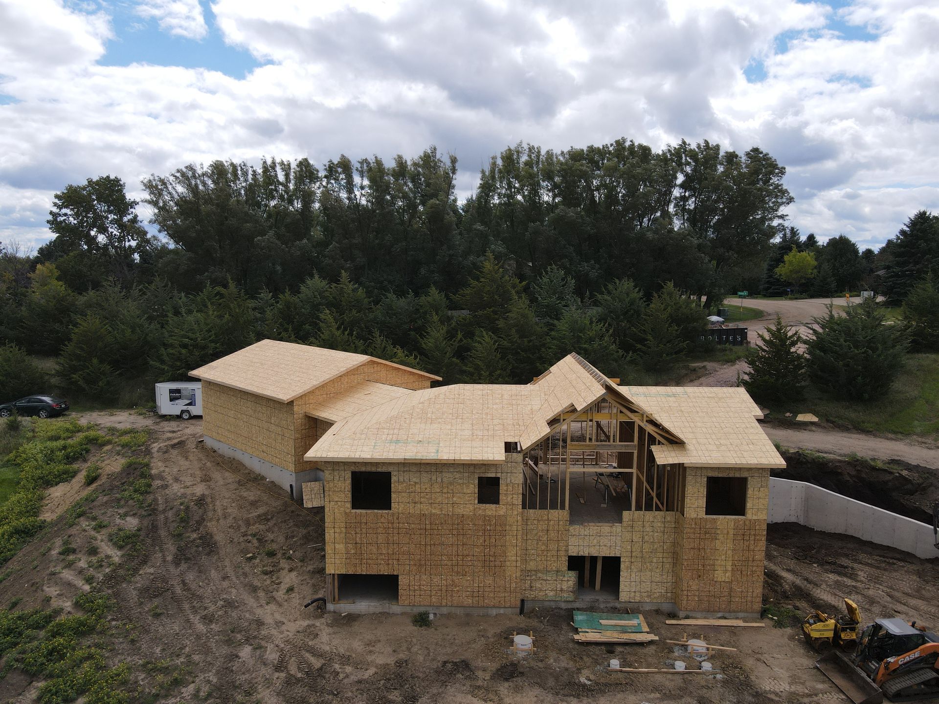 House under construction, wood framing, roof partially built, surrounded by trees.