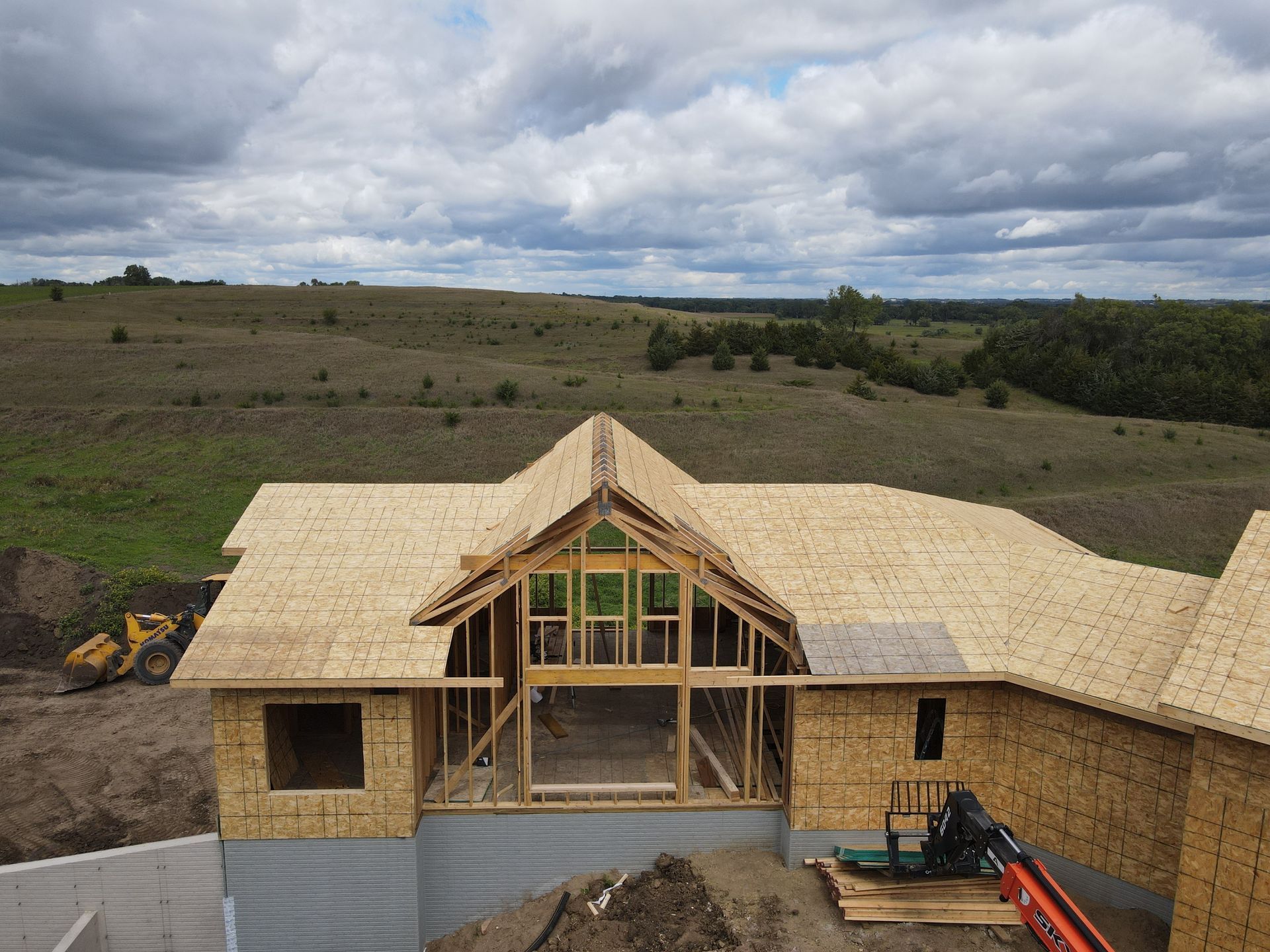 House under construction with exposed framing and sheathing, set against a rolling hillside under a cloudy sky.