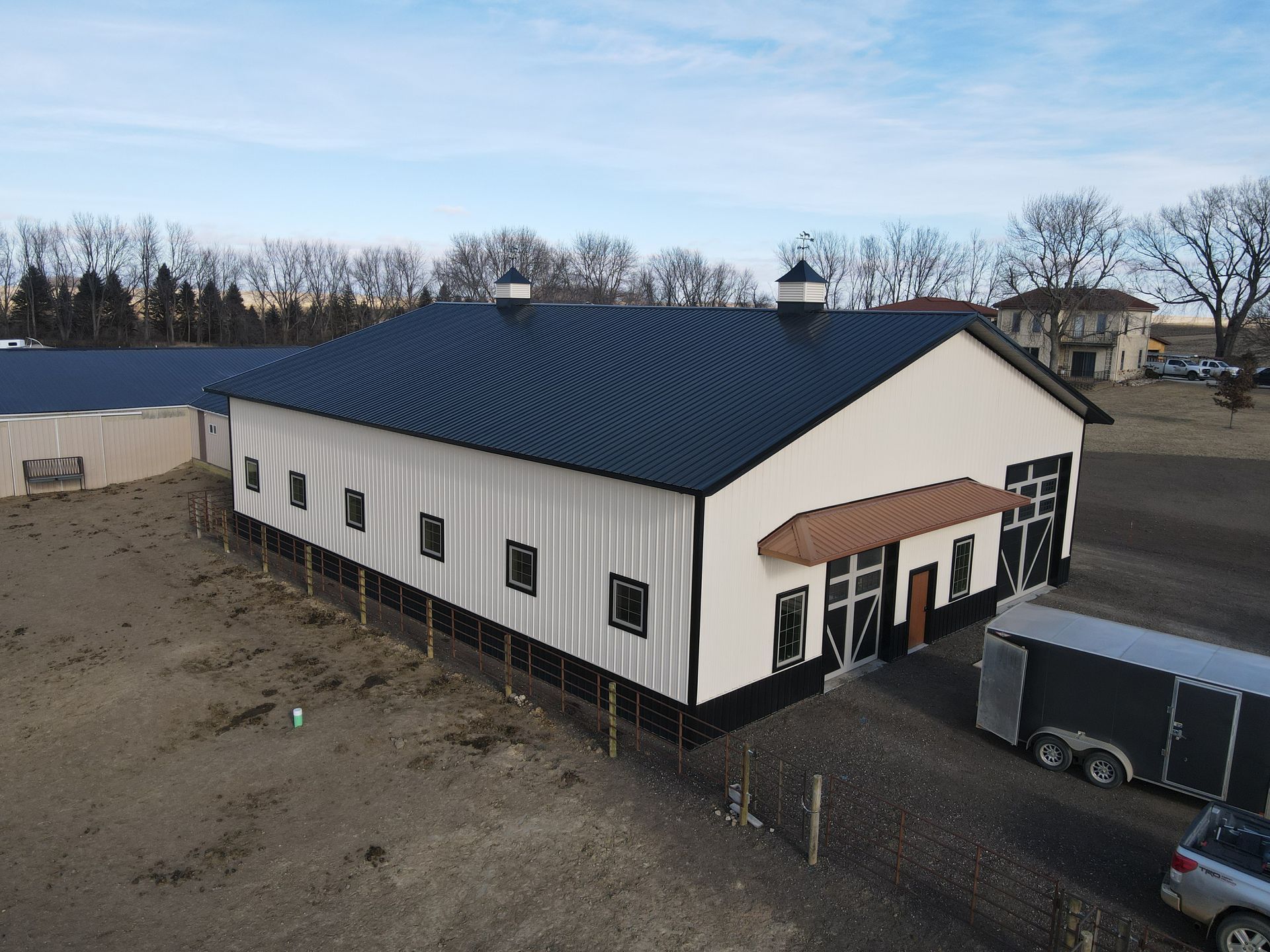 Large white and black barn with a black roof; a horse trailer and car parked in front.