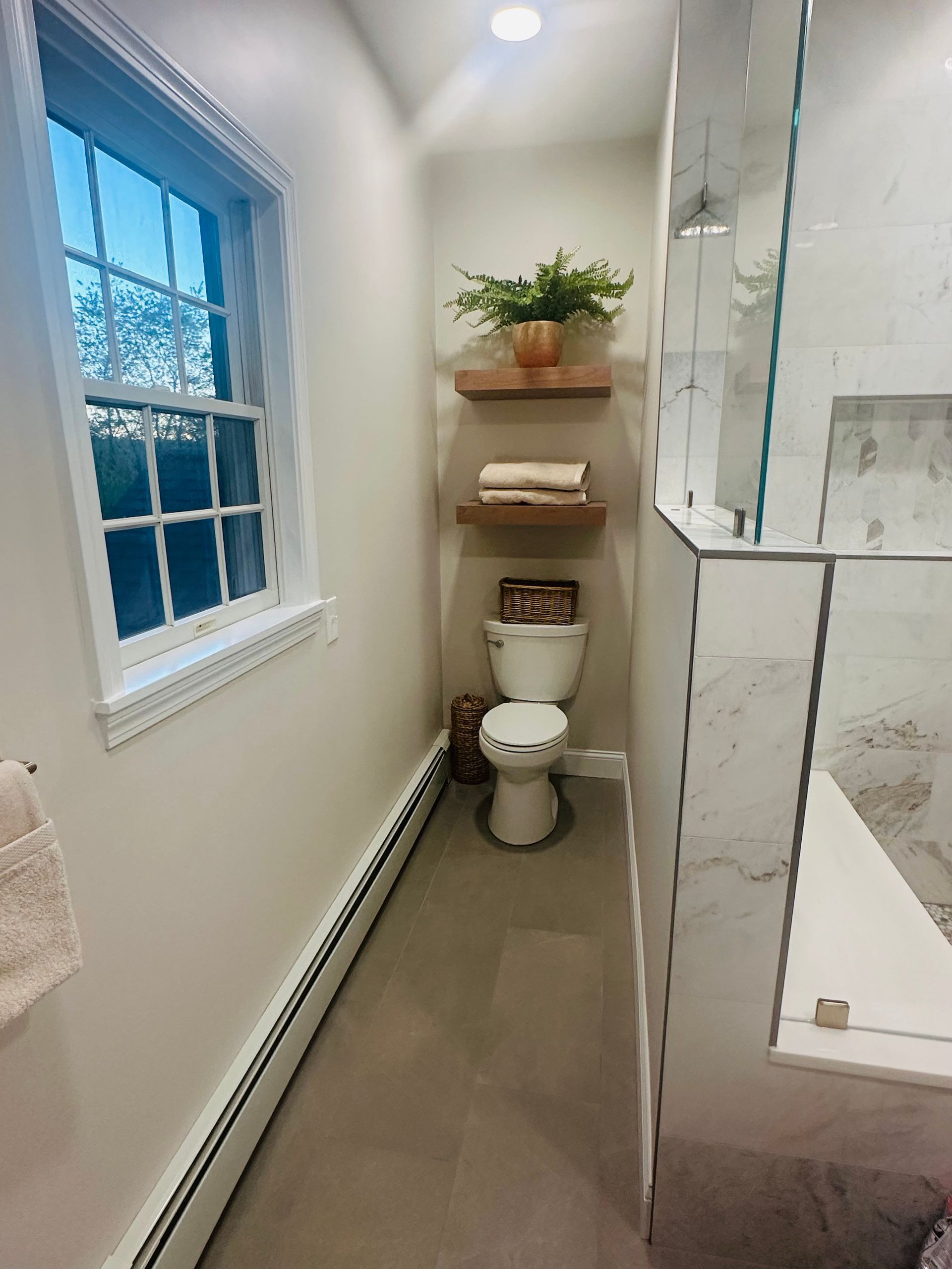 Narrow bathroom with white toilet, floating shelves, and a window beside a glass shower enclosure.