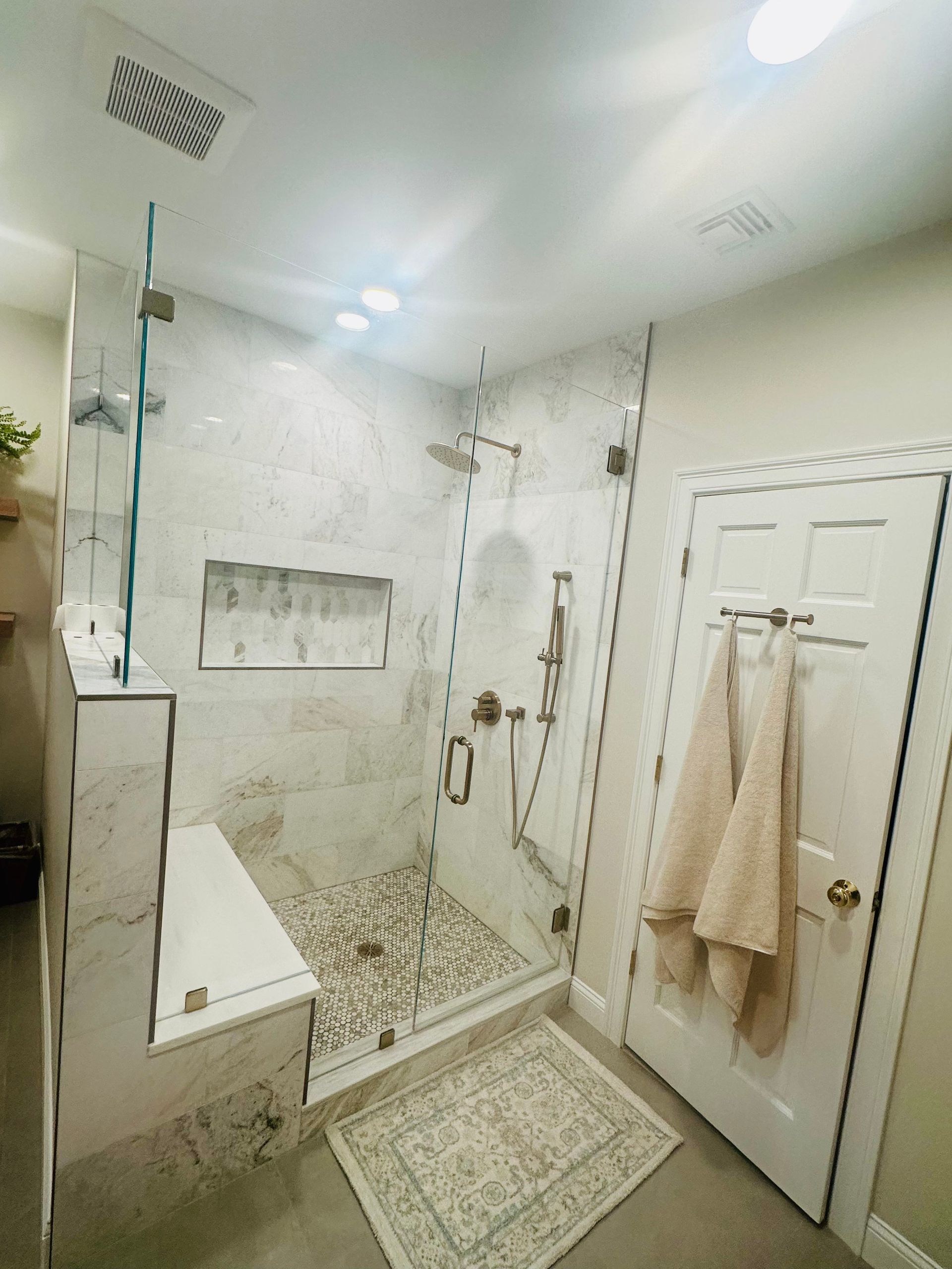 Bright white marble bathroom with glass shower, freestanding tub, vanity, and beige towels on a door.