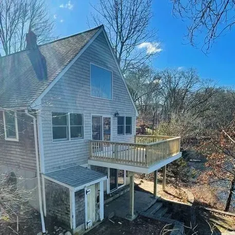 A two-story gray house with a wooden deck overlooks a river on a sunny day.