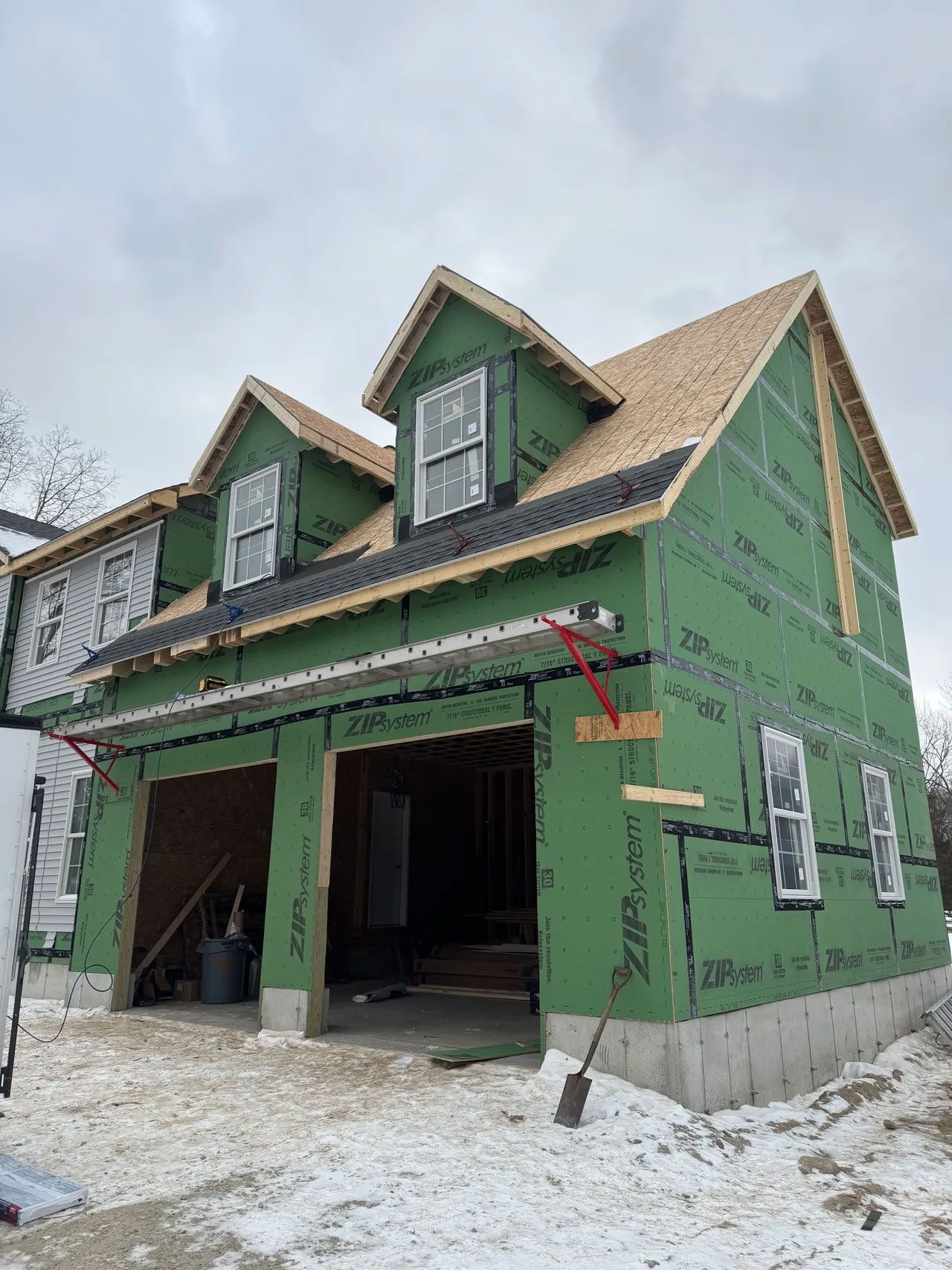 Two-story house under construction, green sheathing, wood framing, two garage doors, dormers, and cloudy sky.