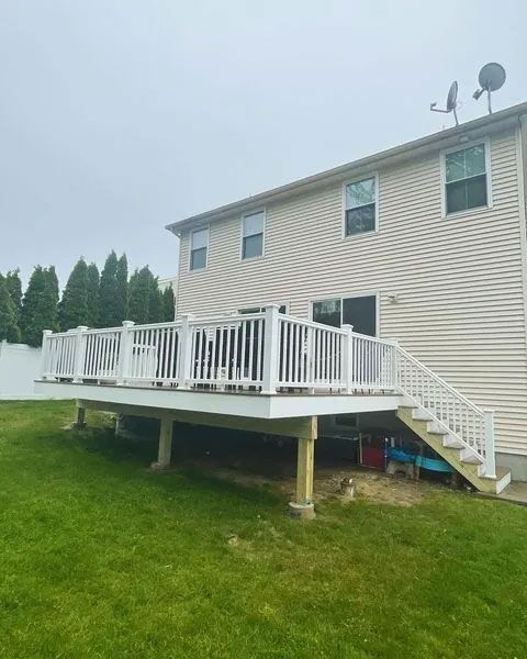 A two-story house with a white deck, stairs, and railing. Green lawn surrounds the deck. Cloudy sky.