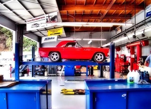 A bright red classic car is raised on a metal lift inside a bright, clean auto repair shop.