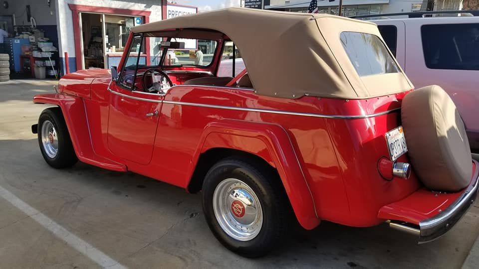 A bright red Willys Jeepster convertible with a tan soft top parked outdoors.