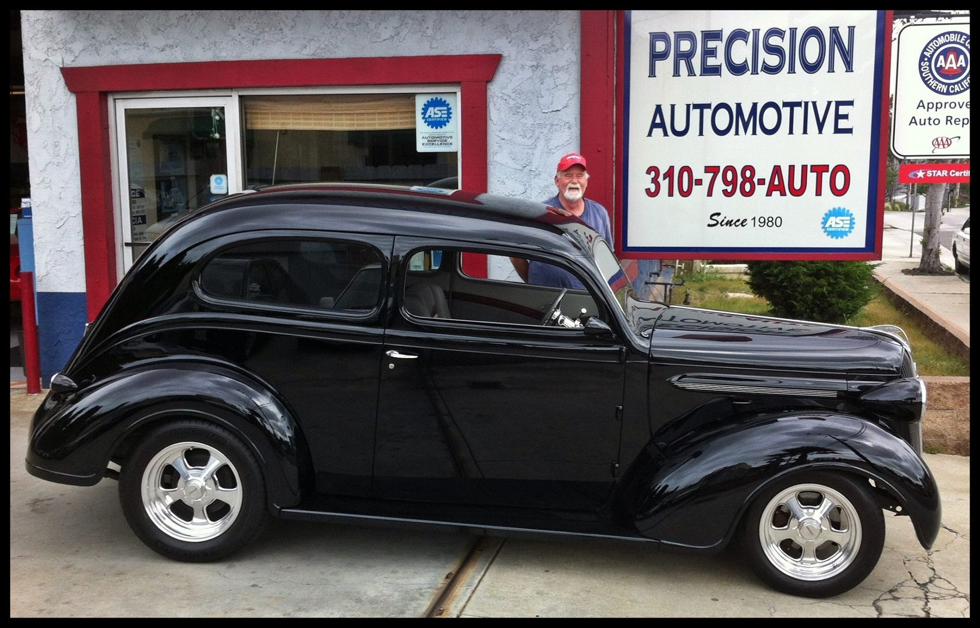 A shiny black vintage car parked in front of Precision Automotive, with a person standing behind it.