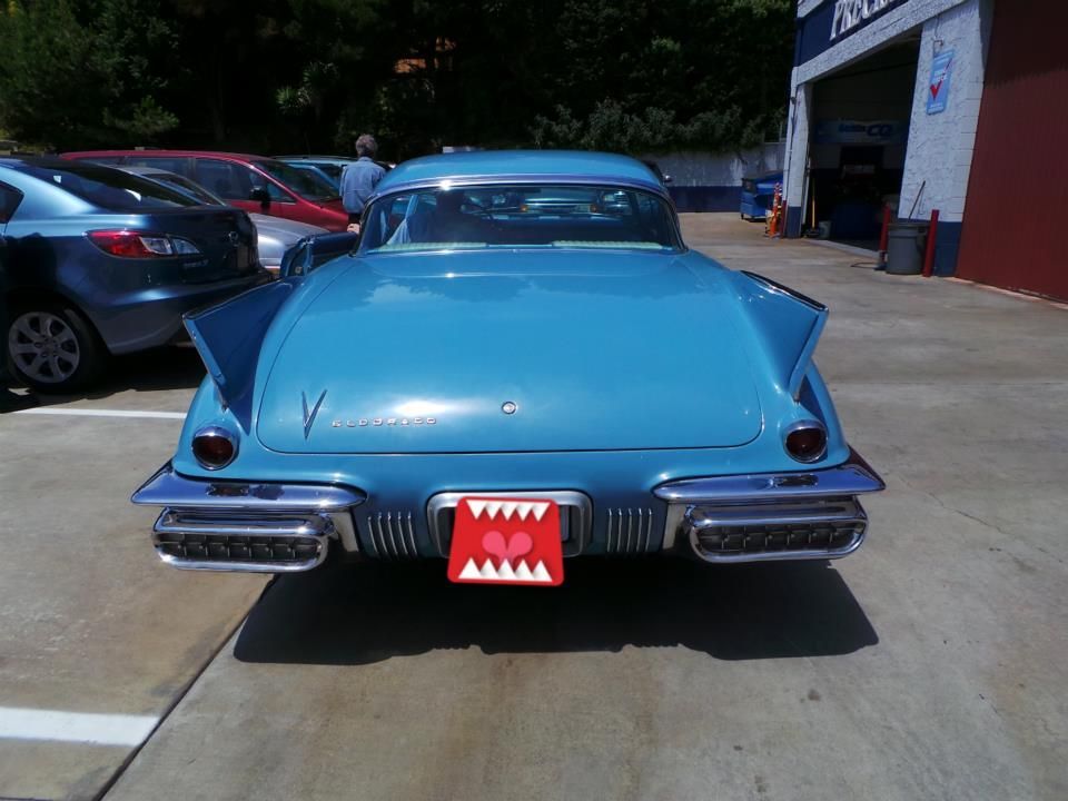 A bright blue vintage car with large fins parked in a concrete lot, with a decorative monster-teeth license plate cover.