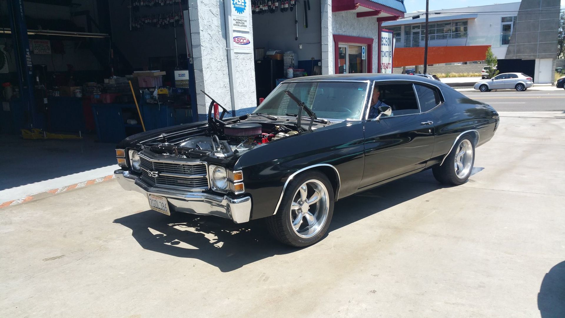 A black vintage Chevrolet Chevelle with its hood open, parked in front of a garage on a sunny day.