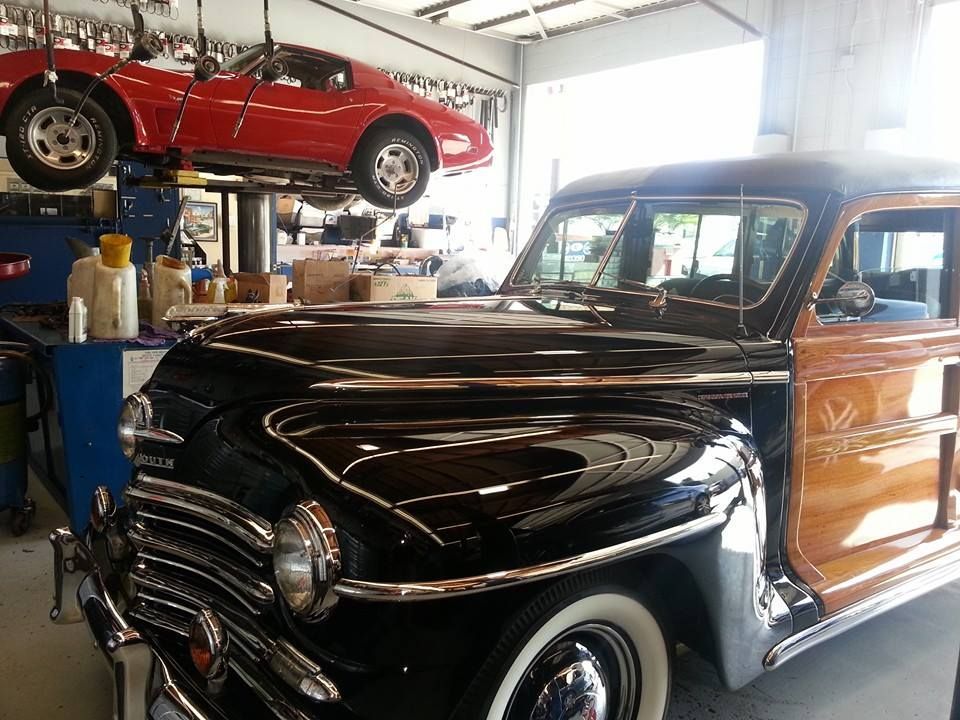 A vintage black and wood-paneled station wagon parked in a garage with a red sports car lifted on a rack above it.