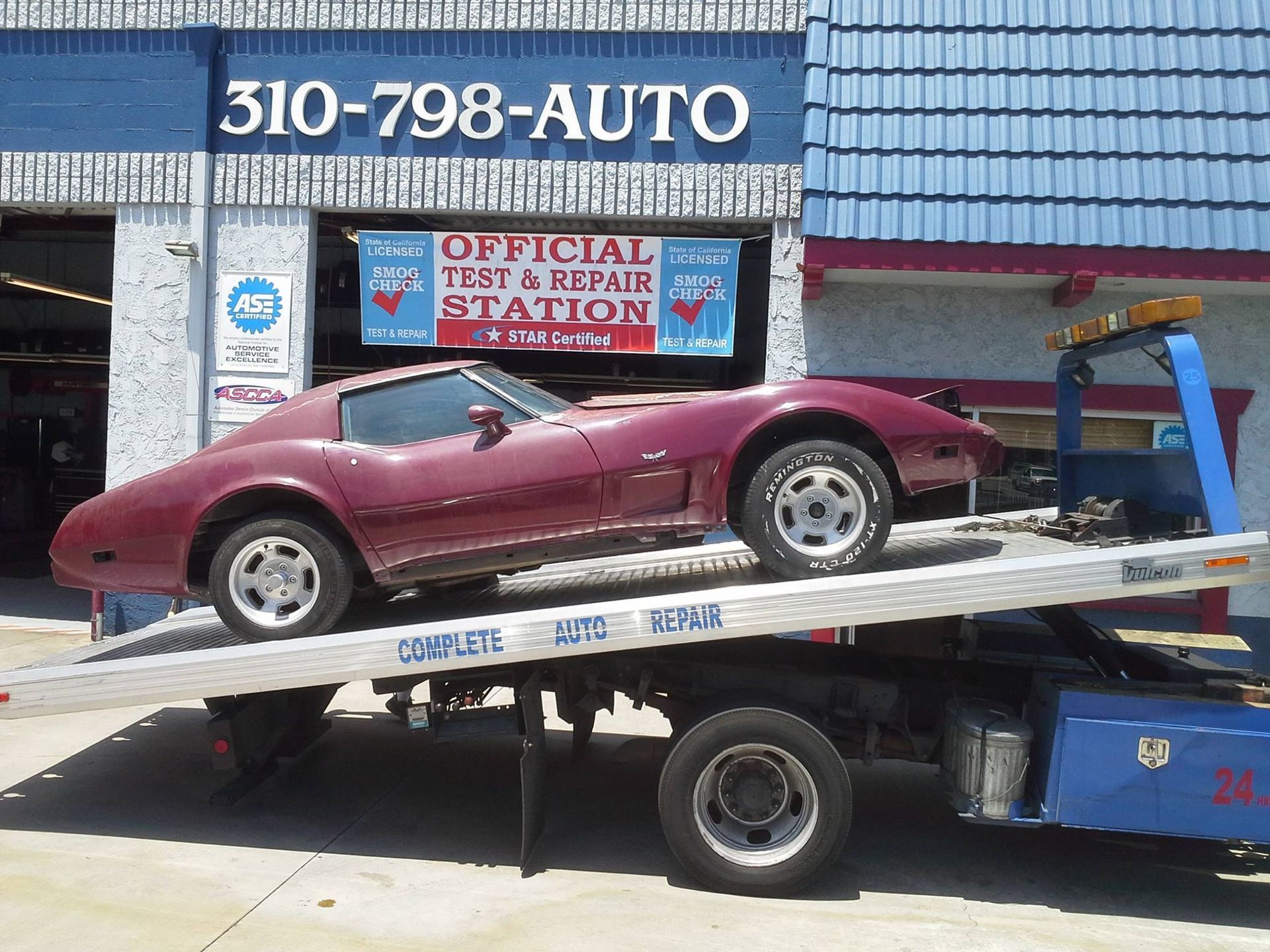 A maroon vintage Corvette sports car sits on a flatbed tow truck in front of an auto repair shop.